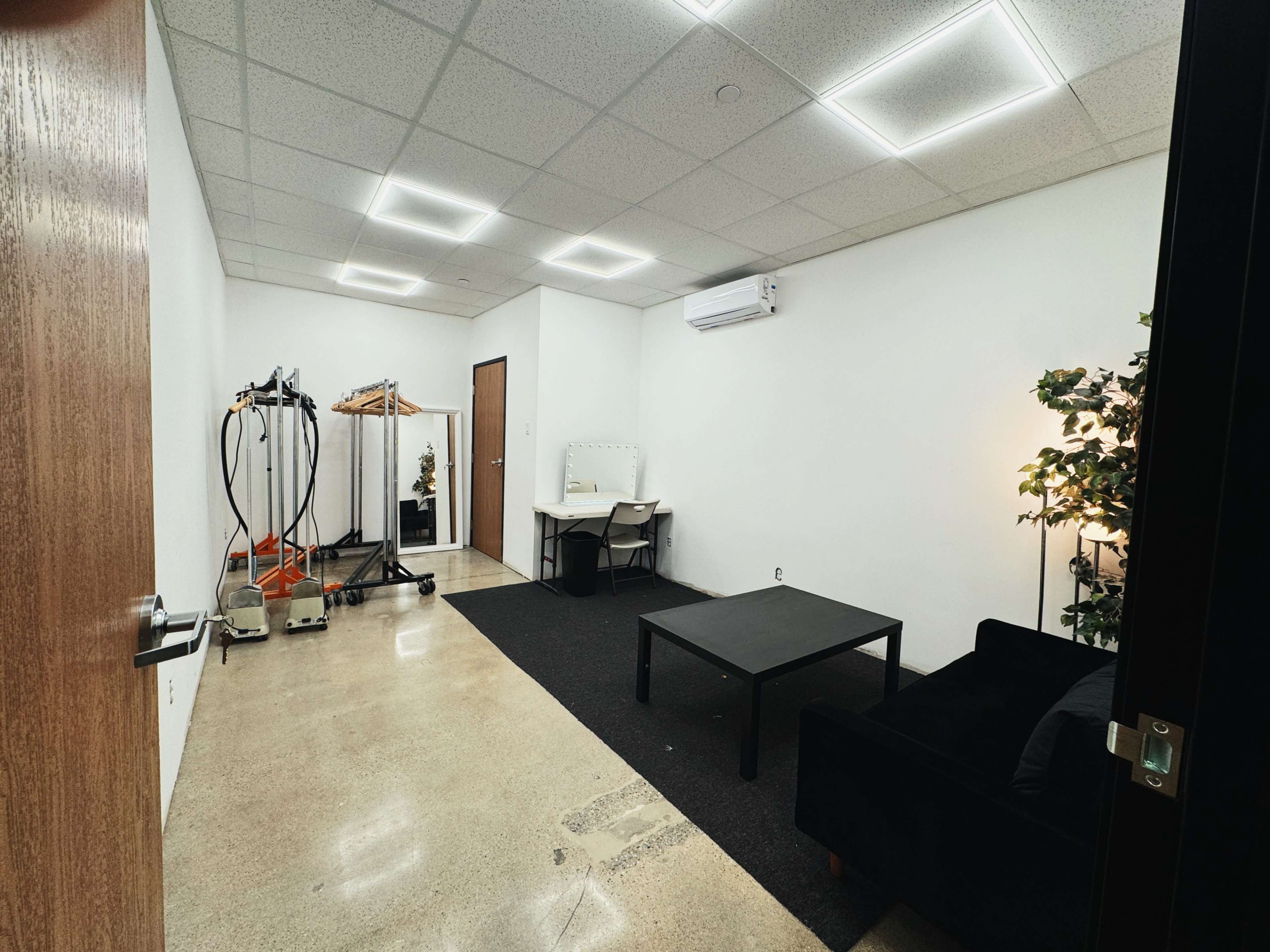 The image shows a sparsely furnished room with a black sofa, a table, a mirror, a clothing rack, and a plant.