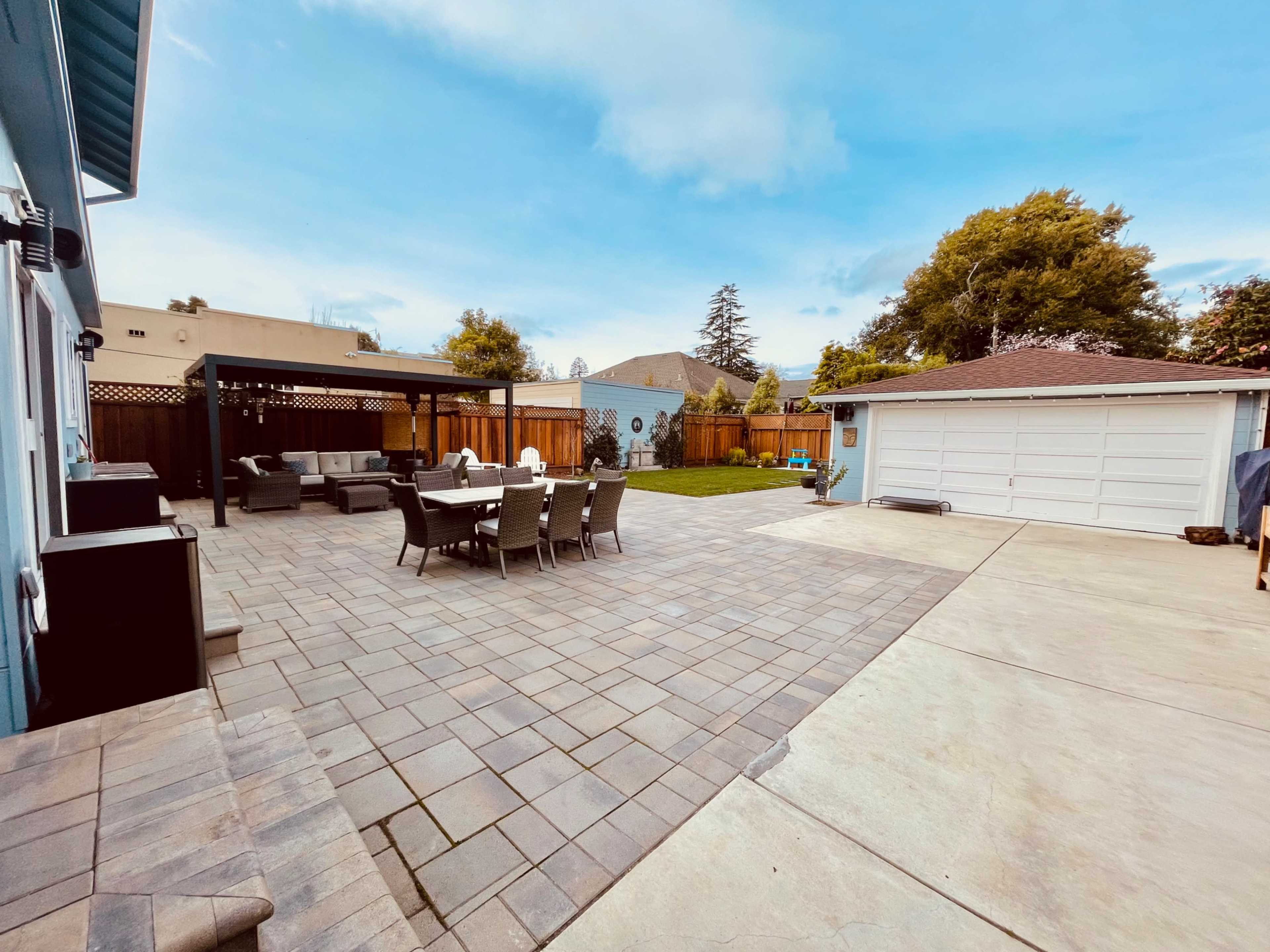 A spacious backyard features a paved patio with a dining set and lounge area, bordered by grass and a garage.