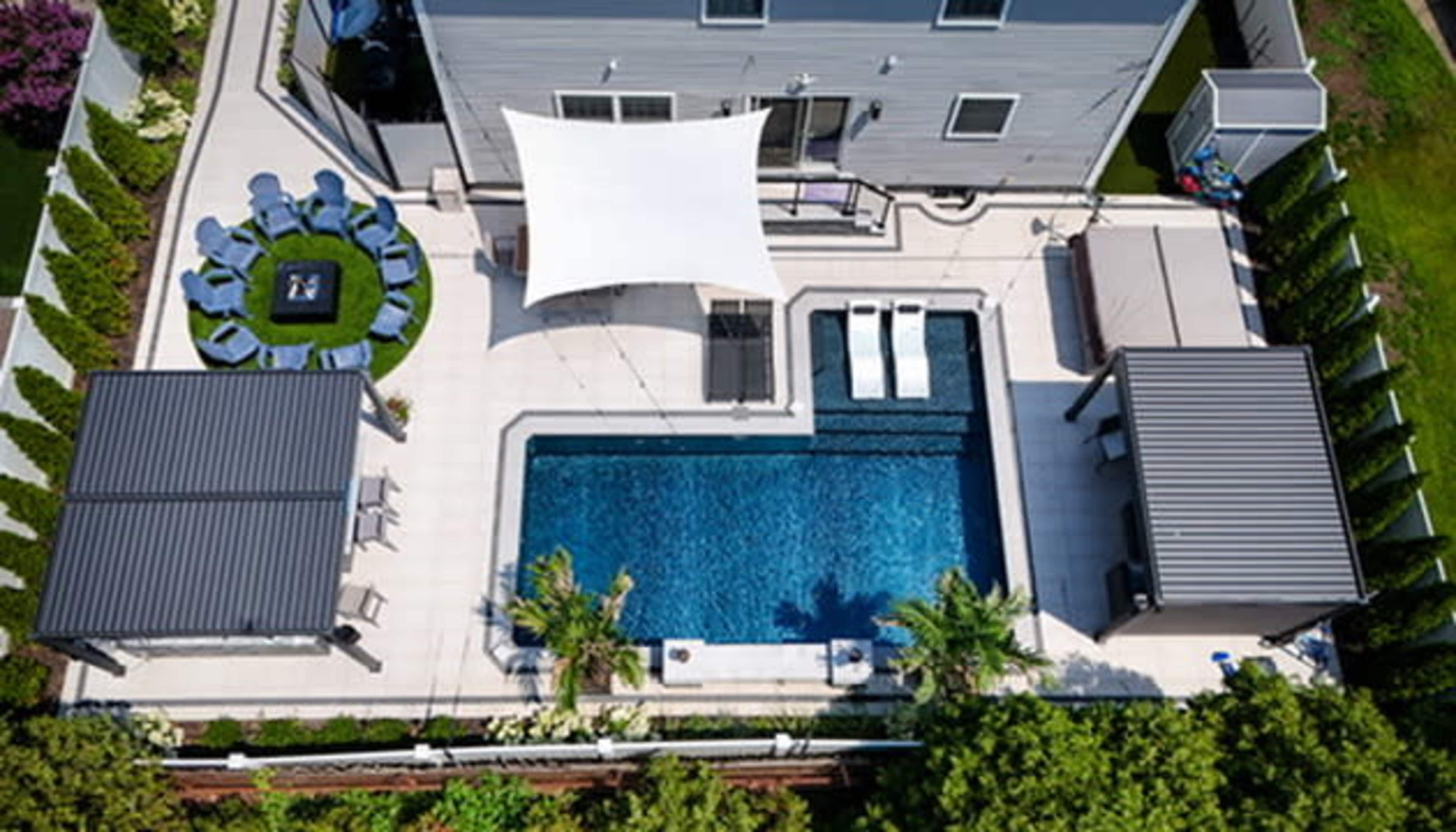 The image features a spacious backyard with a swimming pool surrounded by lounge areas, two shaded pergolas, and neatly arranged seating.