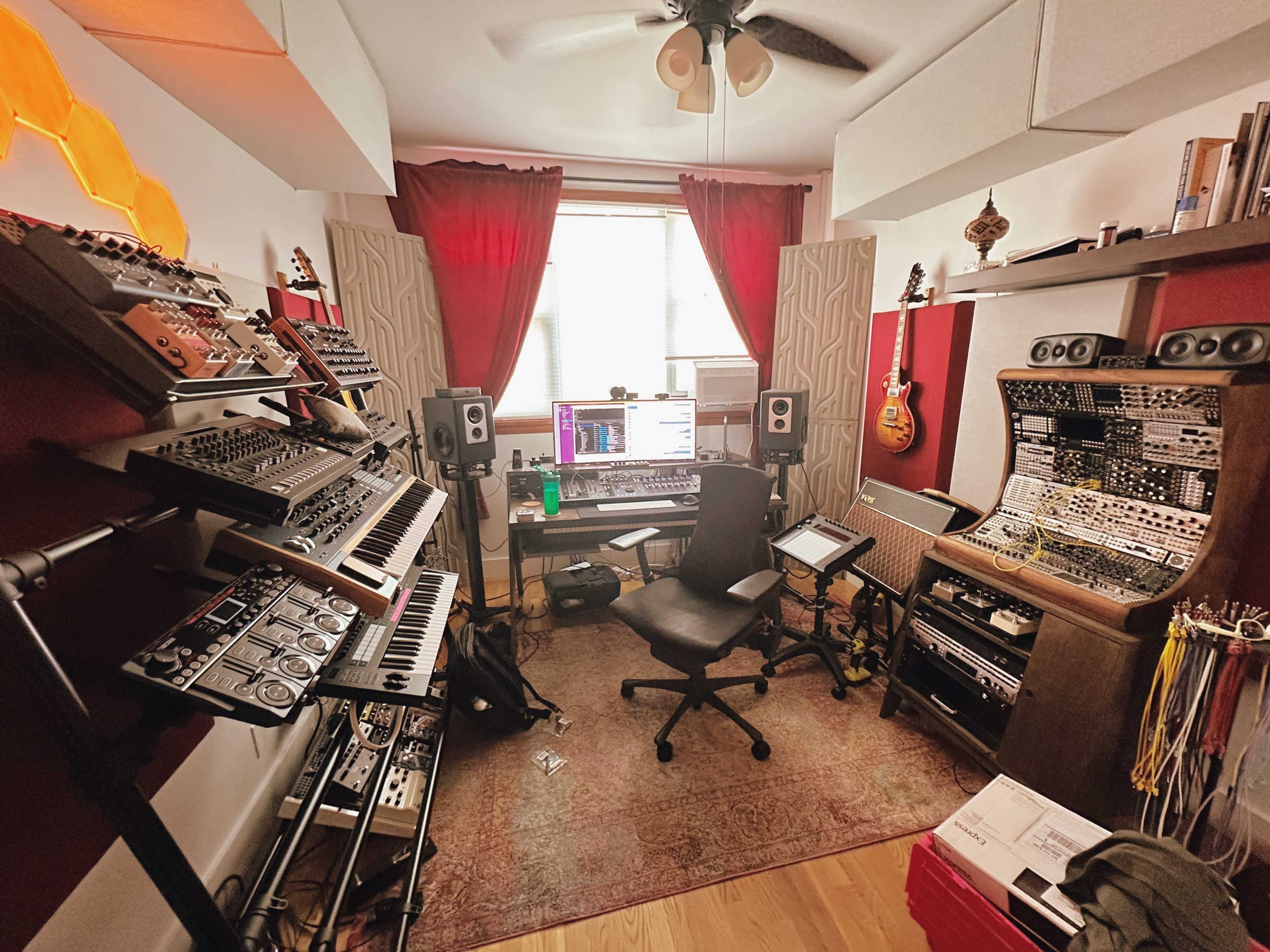 The image shows a compact music studio filled with various synthesizers, monitors, and recording equipment, with a desk and chair positioned in the center.