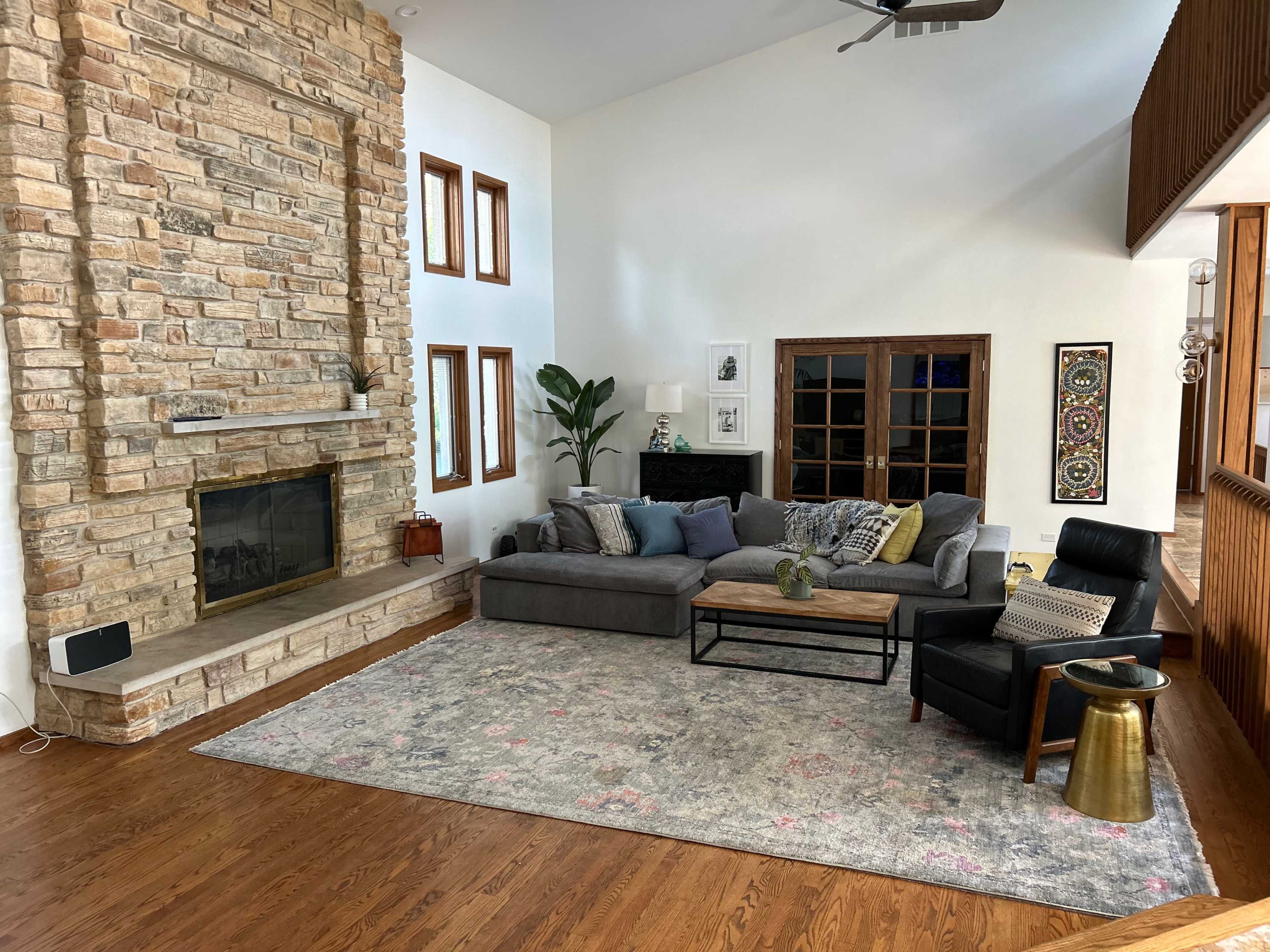A spacious living room featuring a stone fireplace, a gray sectional sofa, a wooden coffee table, and large windows that provide natural light.