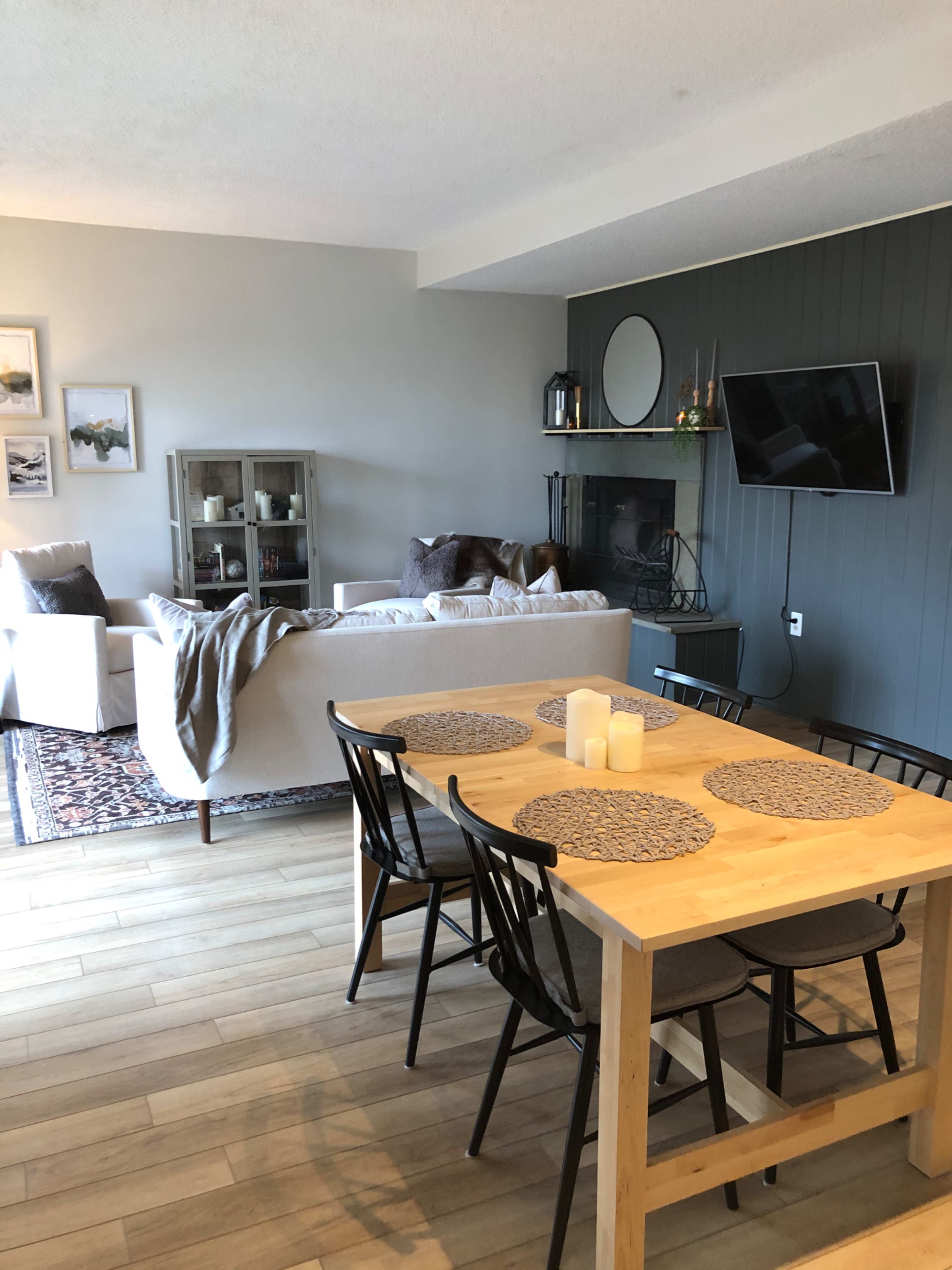A dining area with a wooden table and black chairs is set next to a living space featuring a sofa, a coffee table, a television, and decorative elements on the walls.