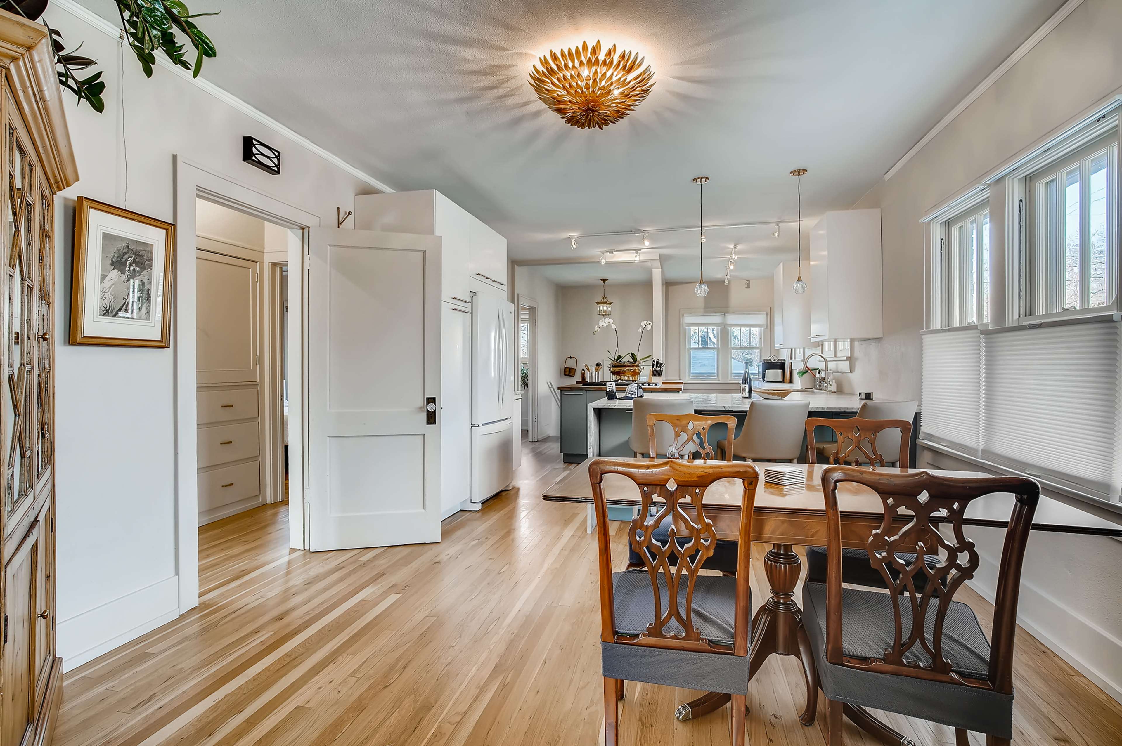 The image shows a bright and open kitchen and dining area with wooden flooring, a centrally placed dining table, and white cabinetry.