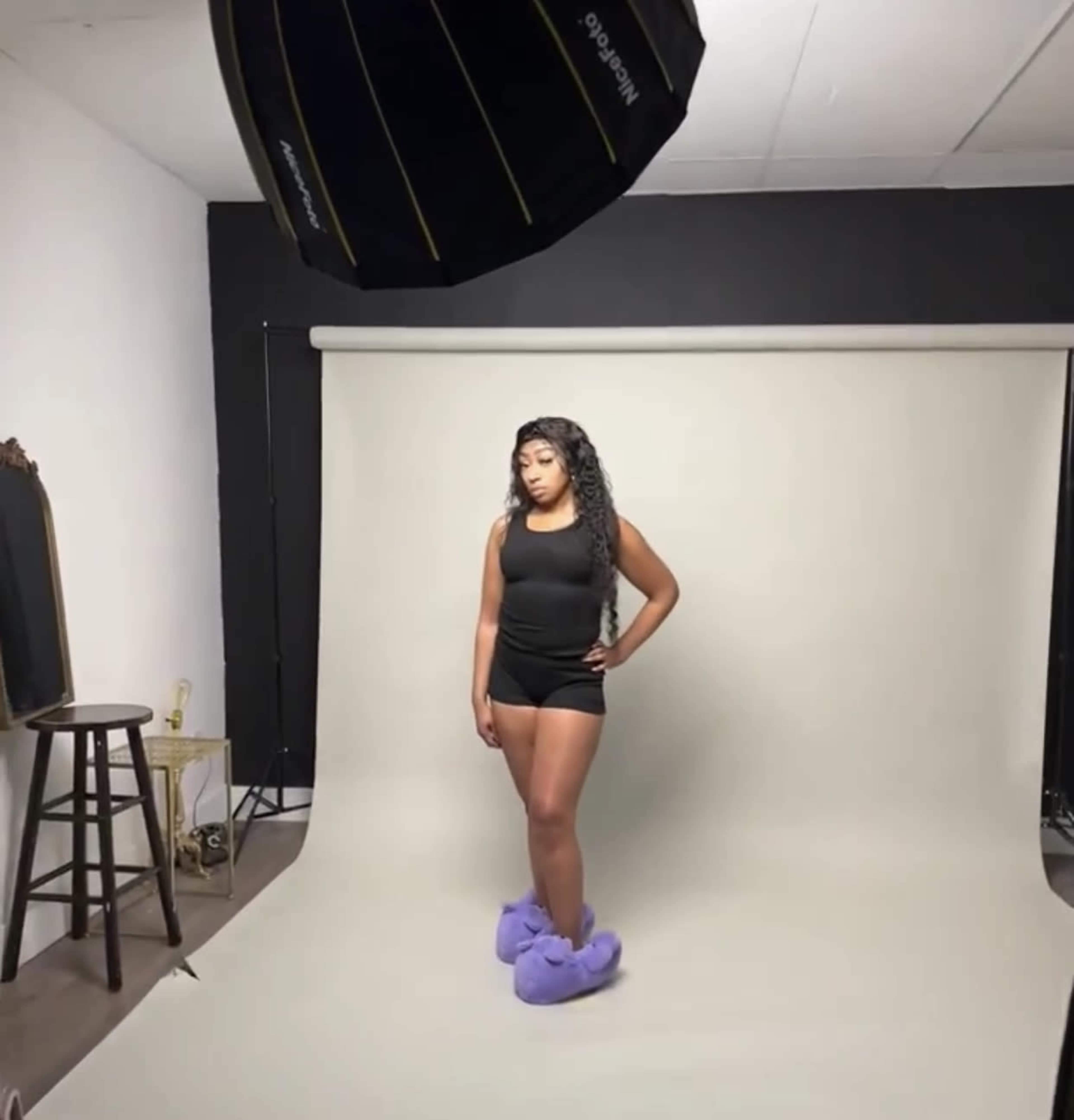 A person stands on a neutral backdrop in a photography studio, wearing a black outfit and large purple slippers.