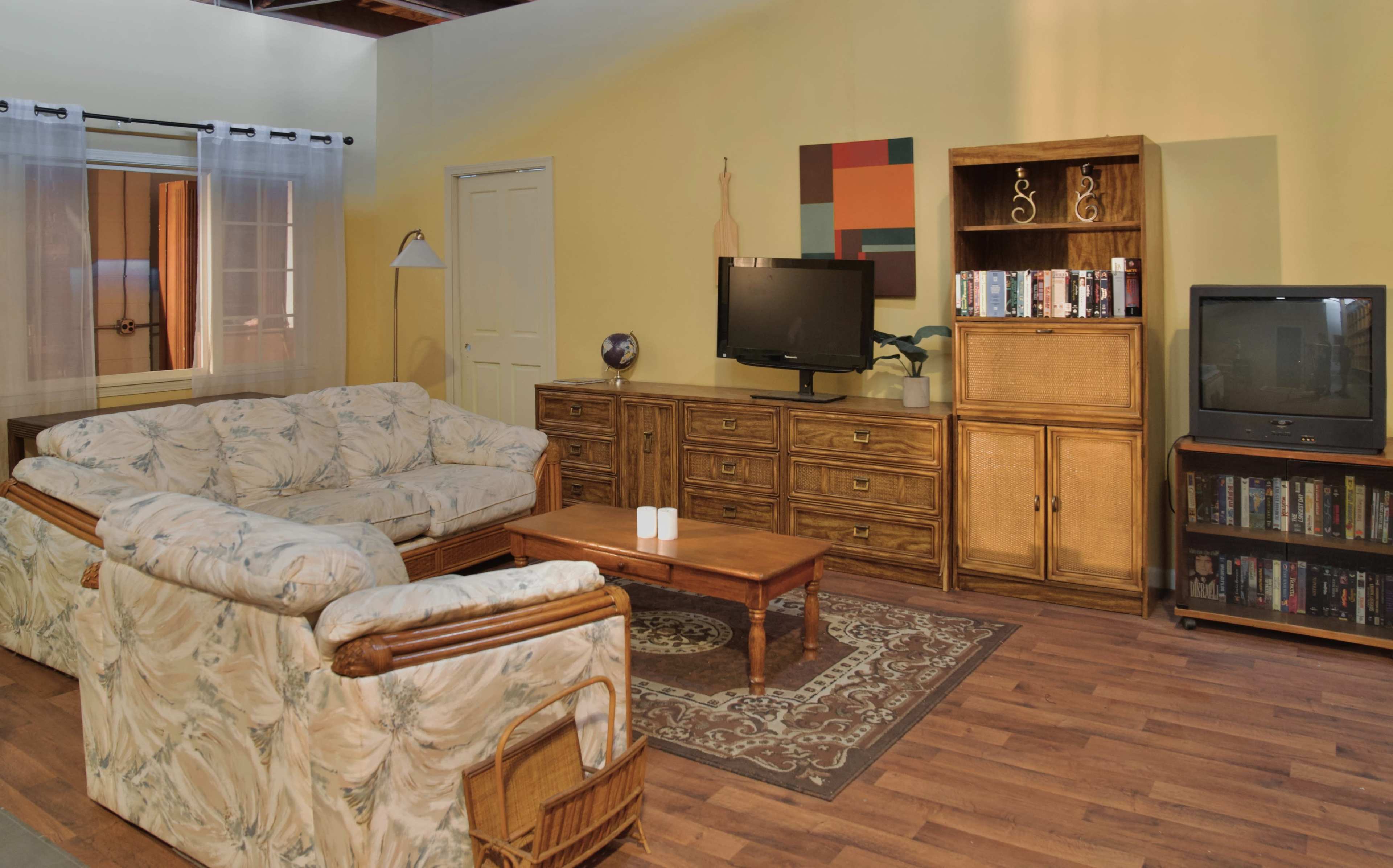 A living room featuring a floral-patterned sofa set, a wooden coffee table, a television on a cabinet, and a bookshelf against a yellow wall.