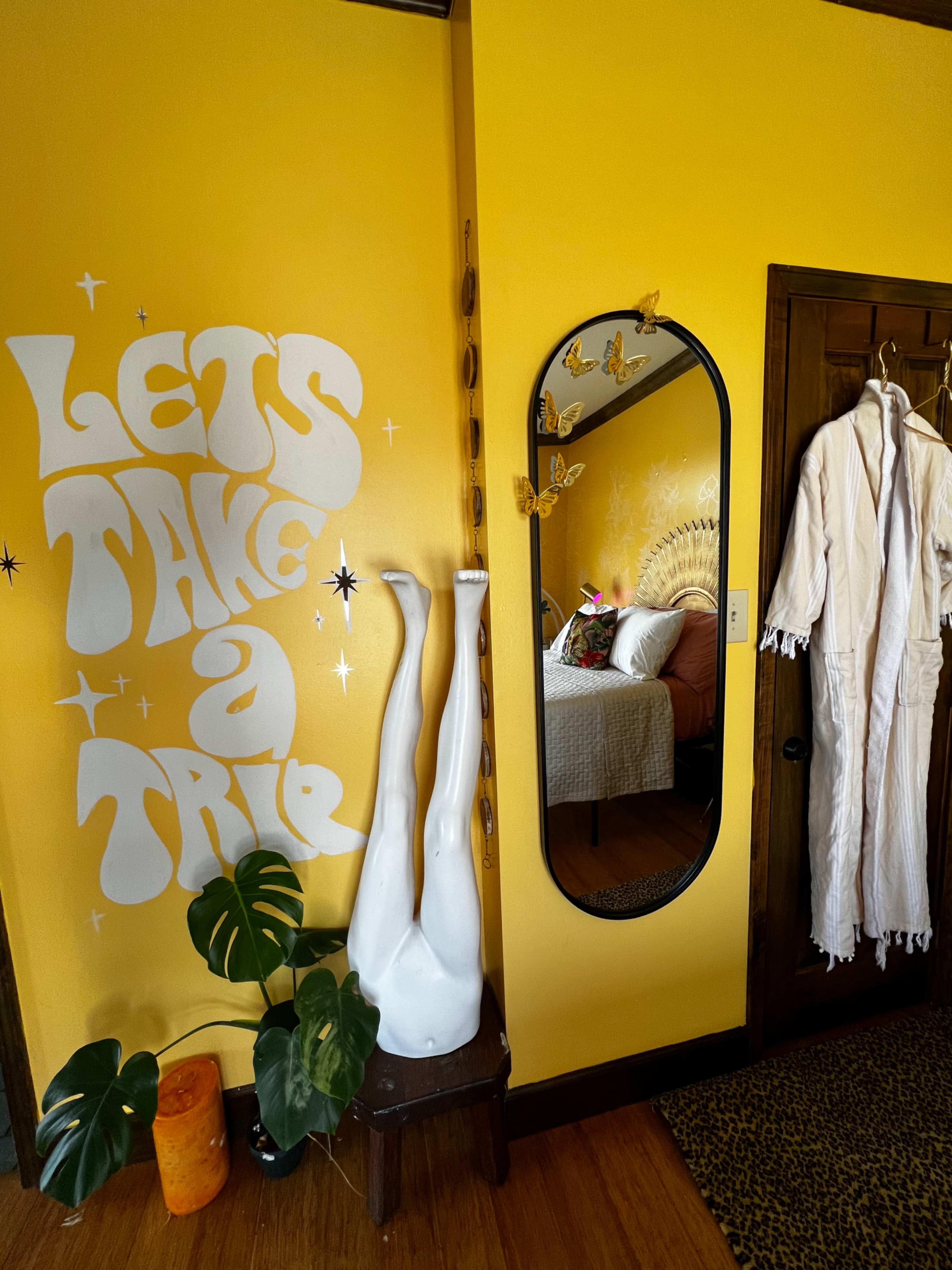 A bright yellow wall with the phrase "LET'S TAKE A TRIP" in white letters, a full-length mirror adorned with butterfly decorations, and a mannequin's legs protruding from a small table beside a green plant and an orange candle.