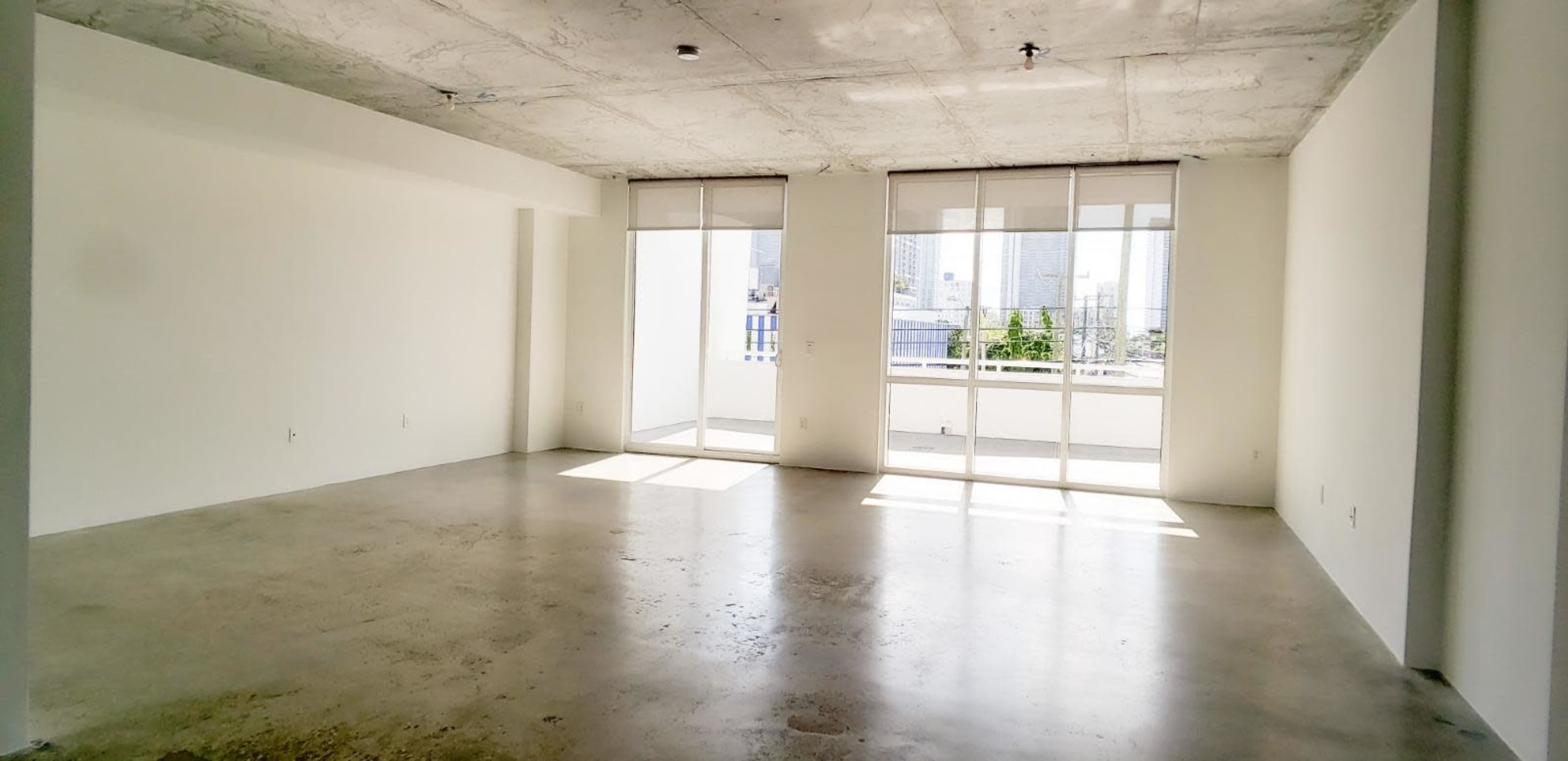The image shows a spacious, empty room with concrete flooring and large windows allowing natural light to enter.