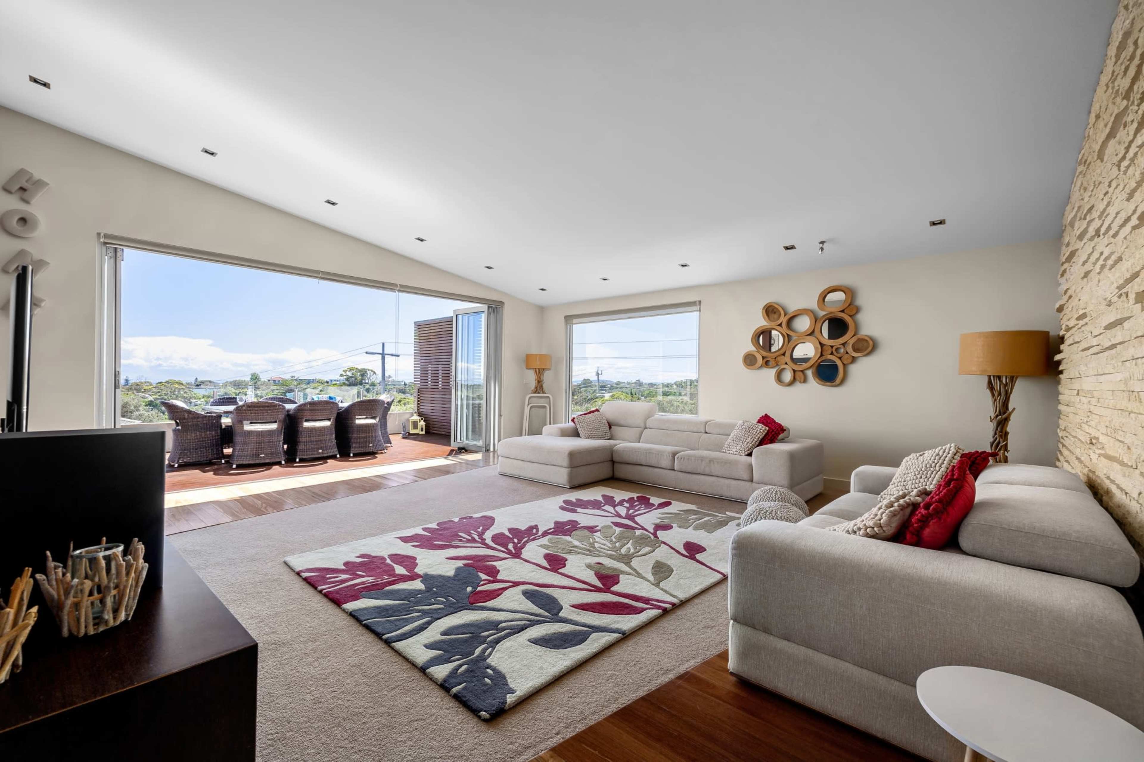 The image shows a modern living room with a sectional sofa, a patterned area rug, large windows opening to an outdoor seating area, and a decorative wall feature.