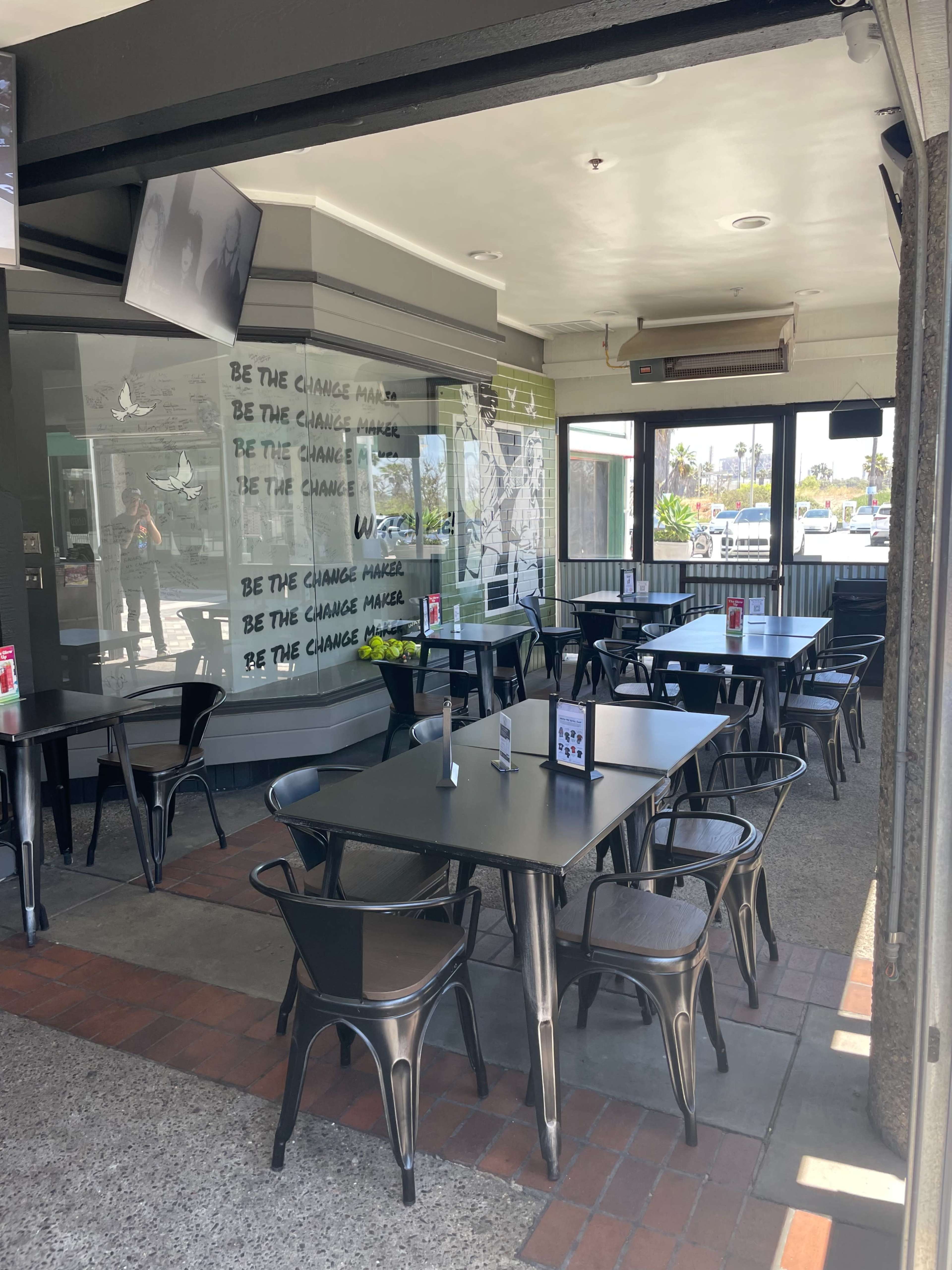 The image shows an empty restaurant patio with black tables and chairs, featuring a glass wall with text and a mural.