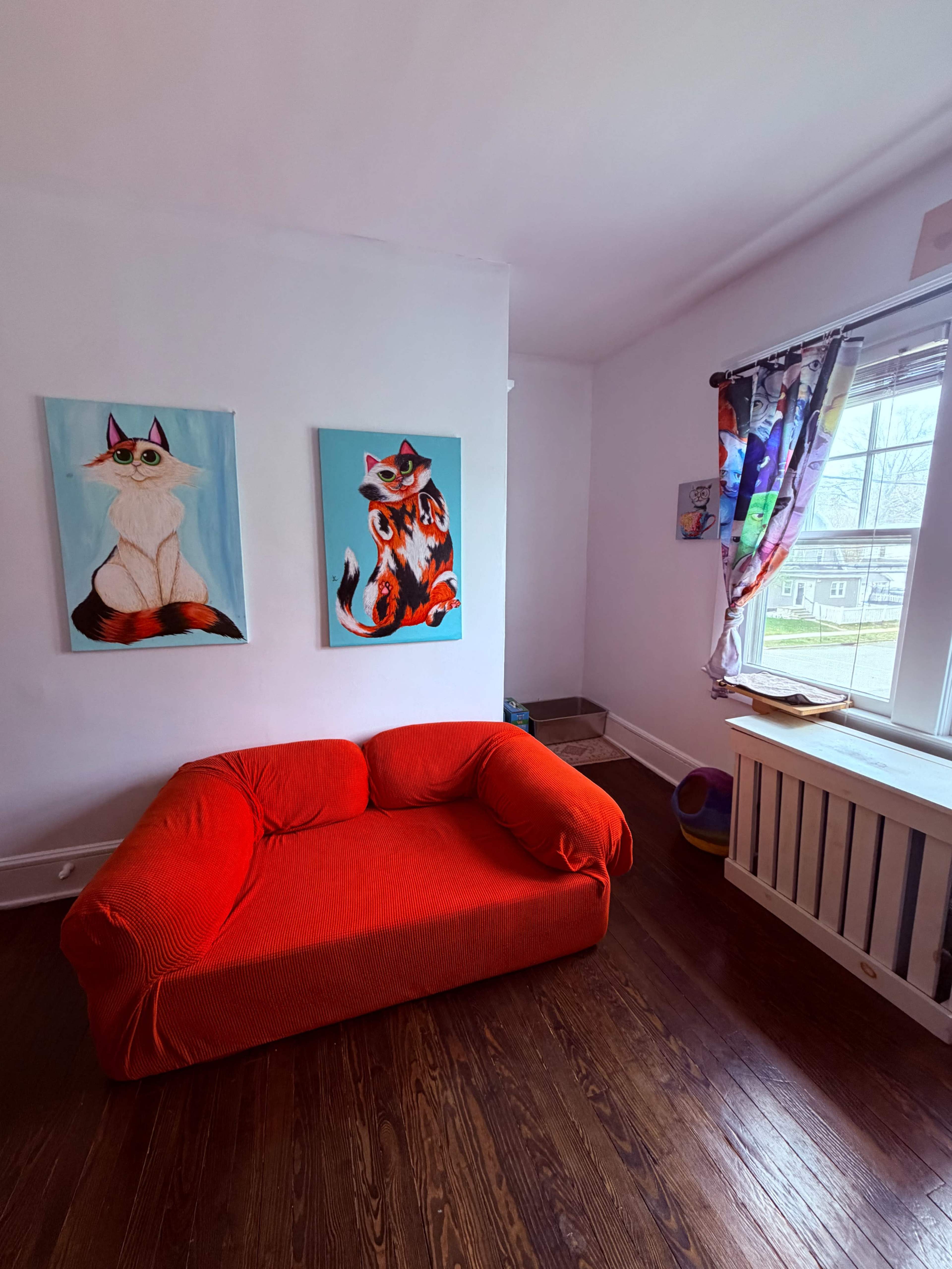 The image shows a small room with a red sofa and two colorful cat paintings on the wall.