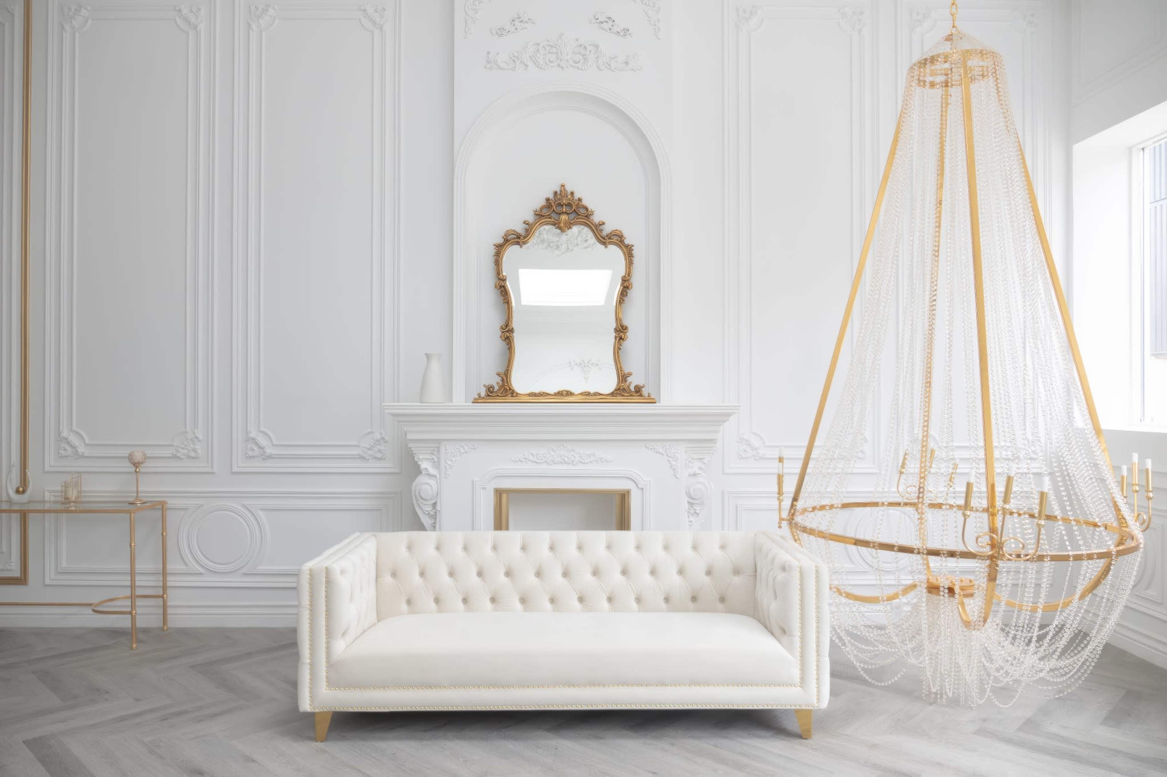 A luxurious white tufted sofa facing a decorative fireplace, with an ornate mirror above it and an elegant chandelier hanging from the ceiling.