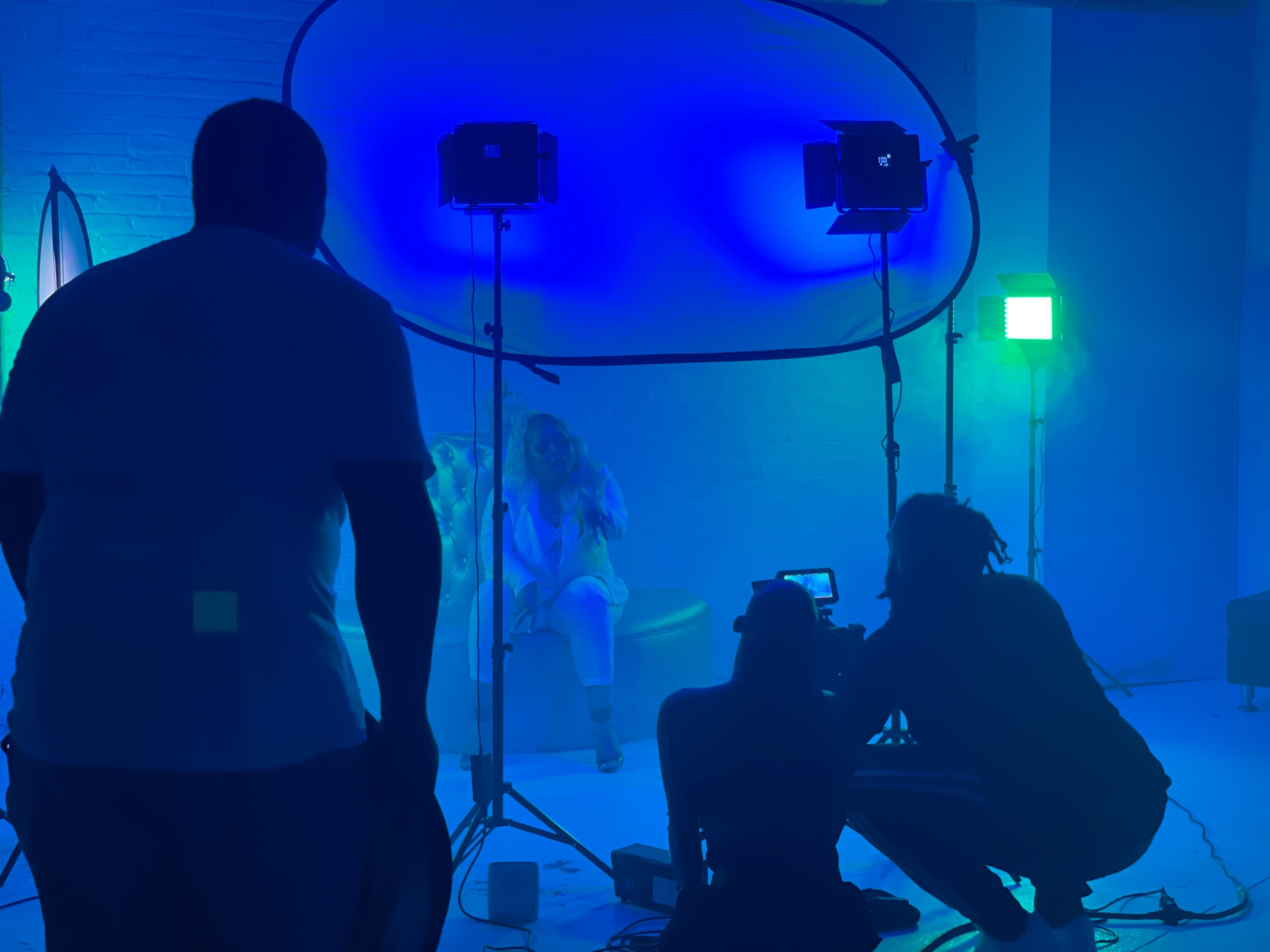 A film crew is set up in a dimly lit studio with blue and green lighting while two individuals are seated in front of the camera.