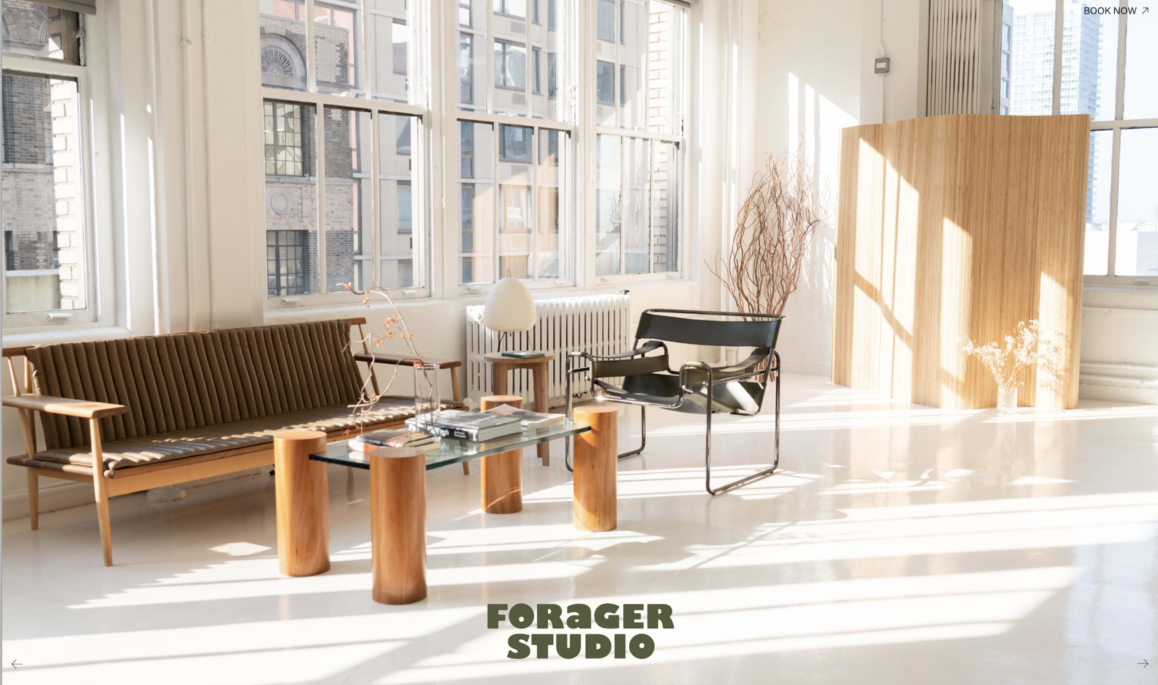 The image shows a bright studio space featuring a wooden sofa, a glass coffee table supported by wooden columns, and a minimalist black chair, with sunlight streaming through large windows.