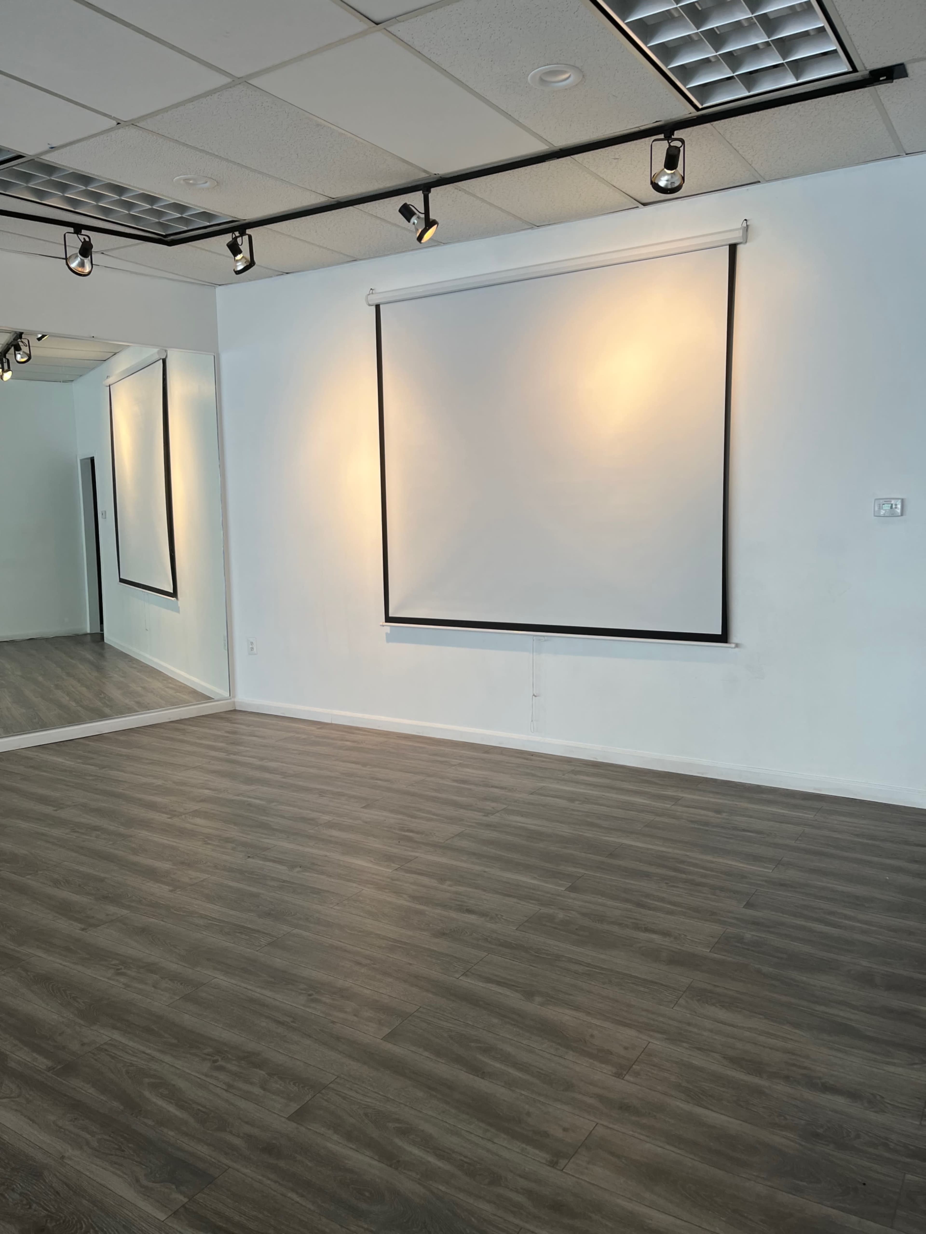 A spacious room with wooden flooring, equipped with a projection screen and a large mirror on one wall.