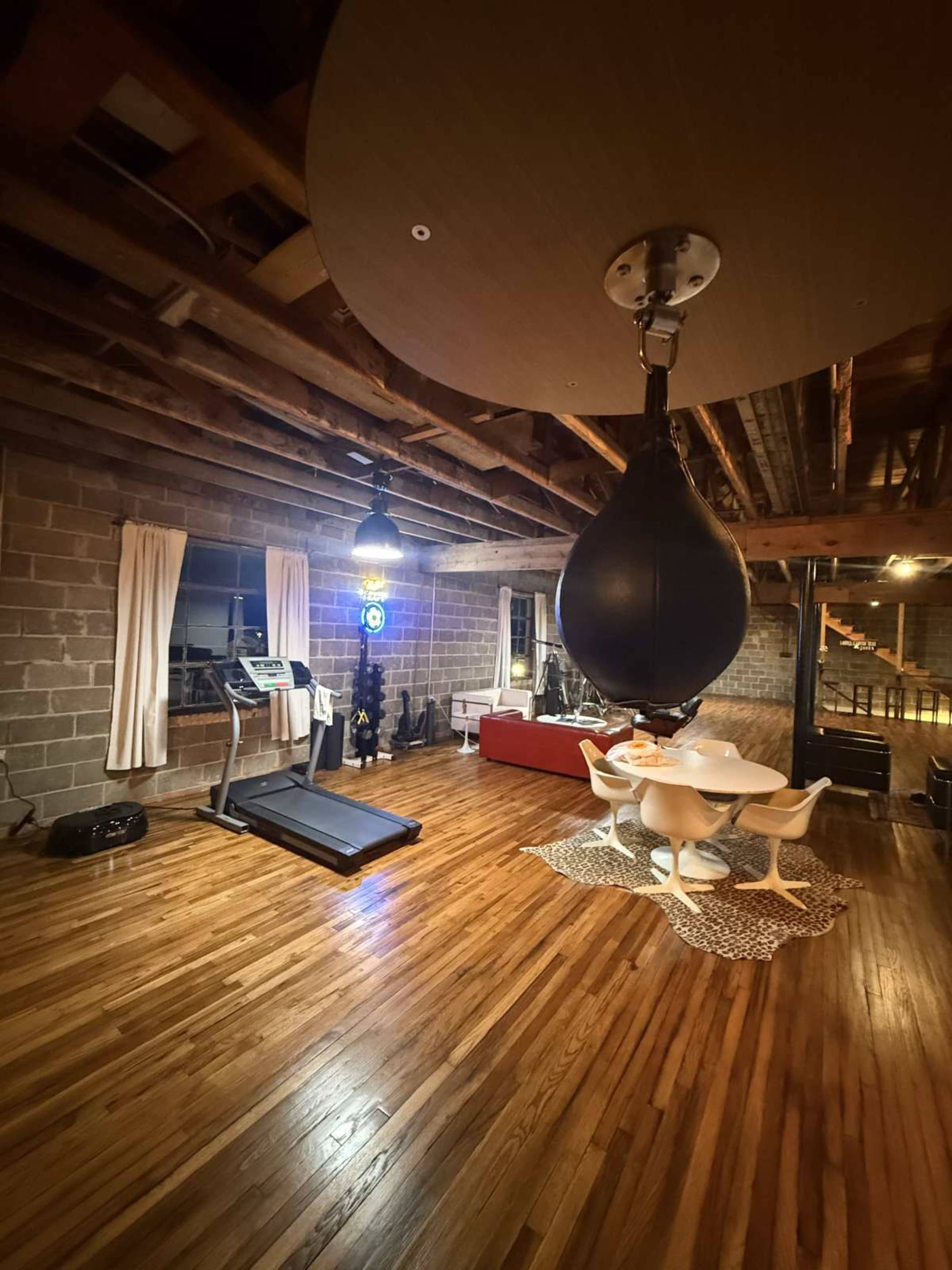 The image shows a spacious room with wooden floors featuring a treadmill, a red couch, a dining table with white chairs, and a hanging punching bag.