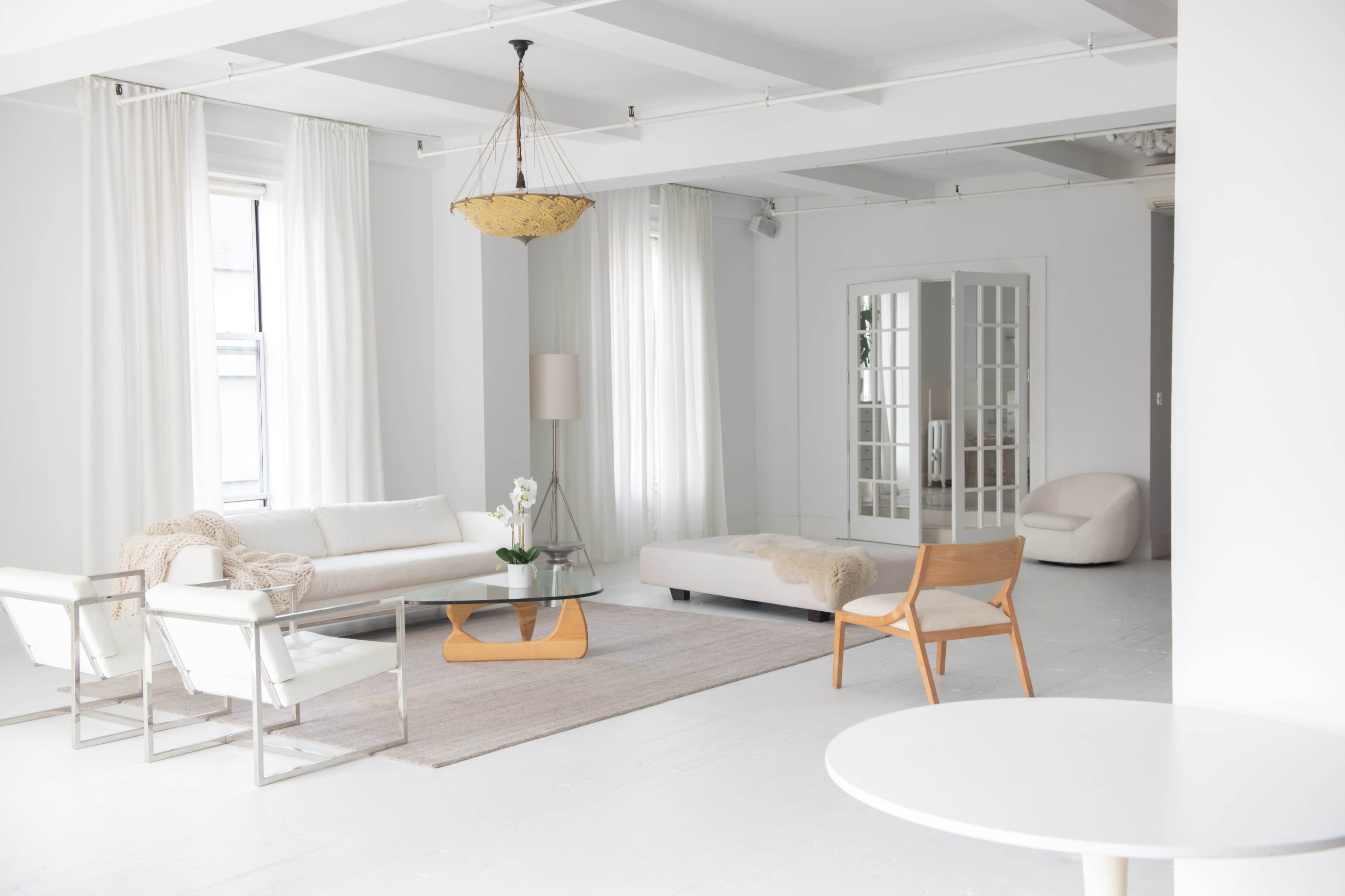 A bright, minimalist living room featuring a white sofa, a glass coffee table, and a light-colored area rug, with large windows and neutral decor.