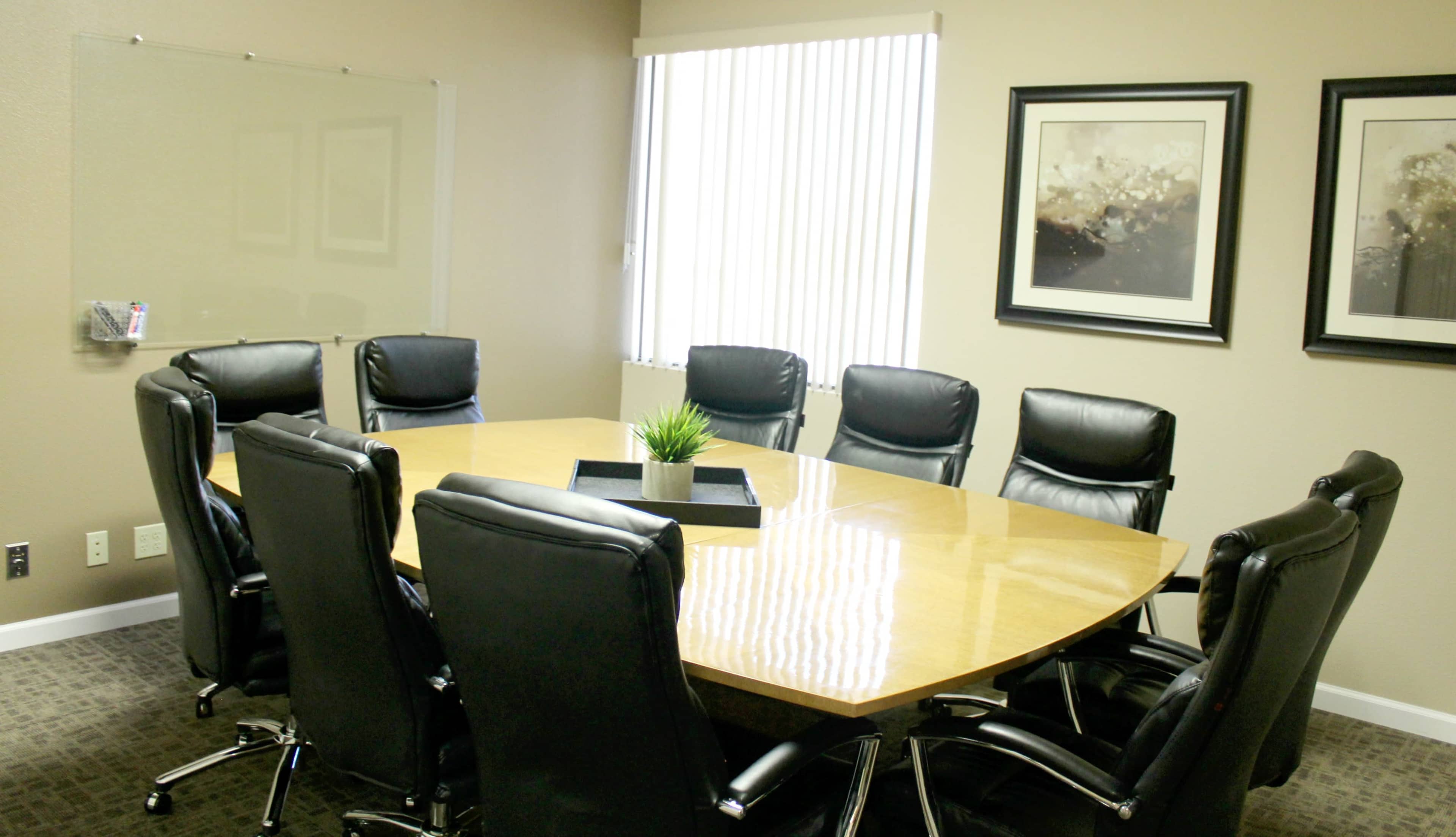 A conference room features a large oval table surrounded by eight black leather chairs, with framed artwork on the walls and a glass board on one side.
