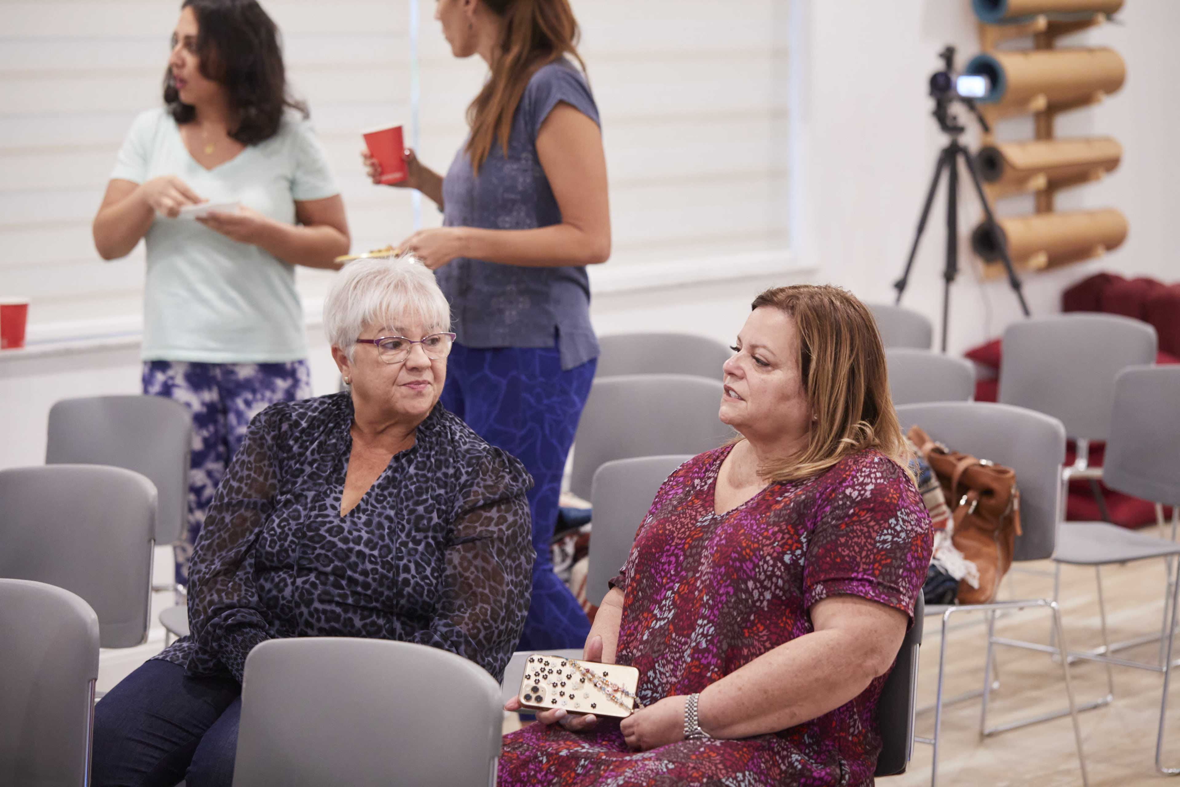 Two women are seated in a room with chairs, while others stand nearby engaged in conversation.