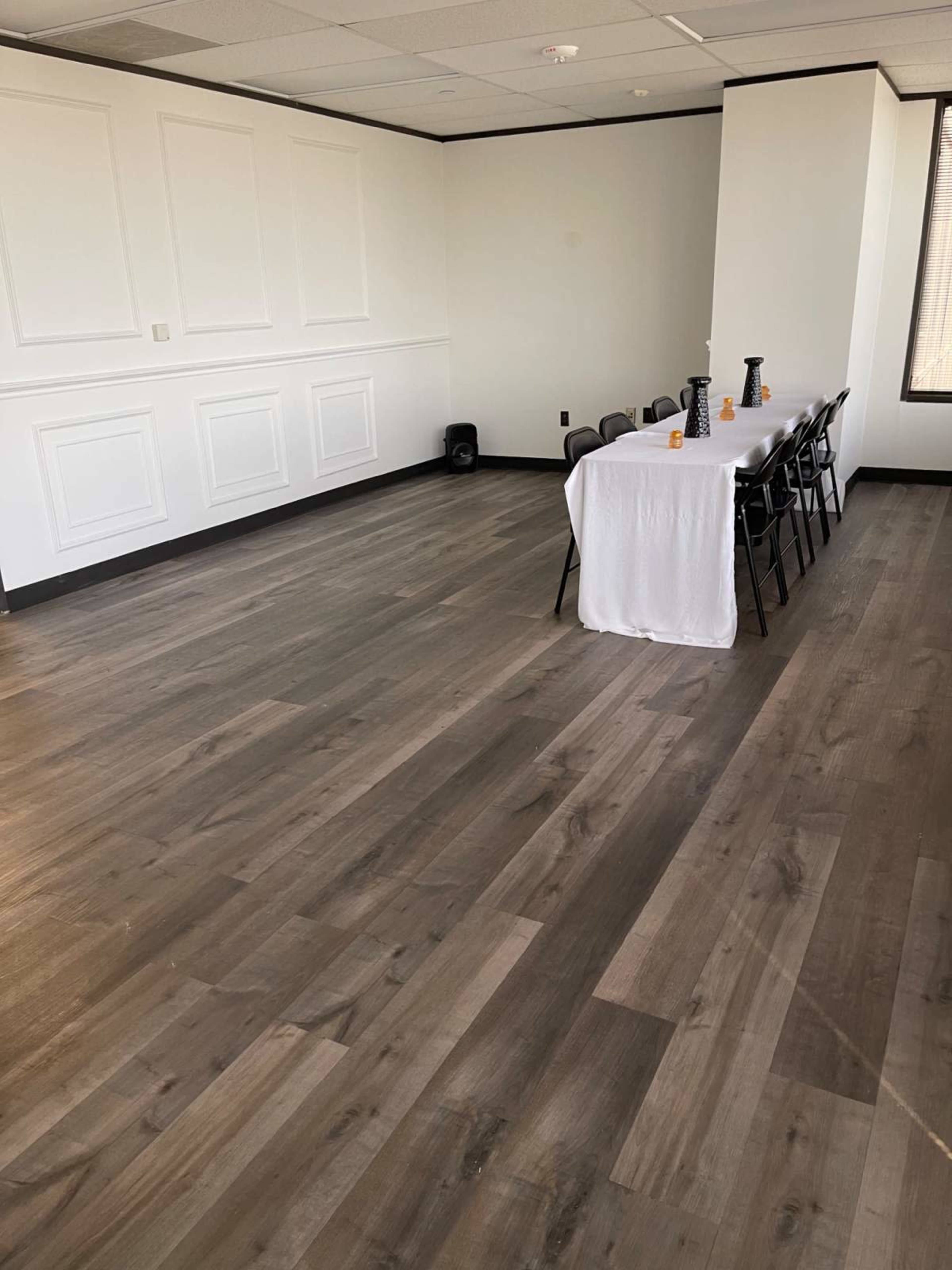 The image shows an empty room with light-colored walls, a long white table set with a tablecloth, and black chairs arranged around it.