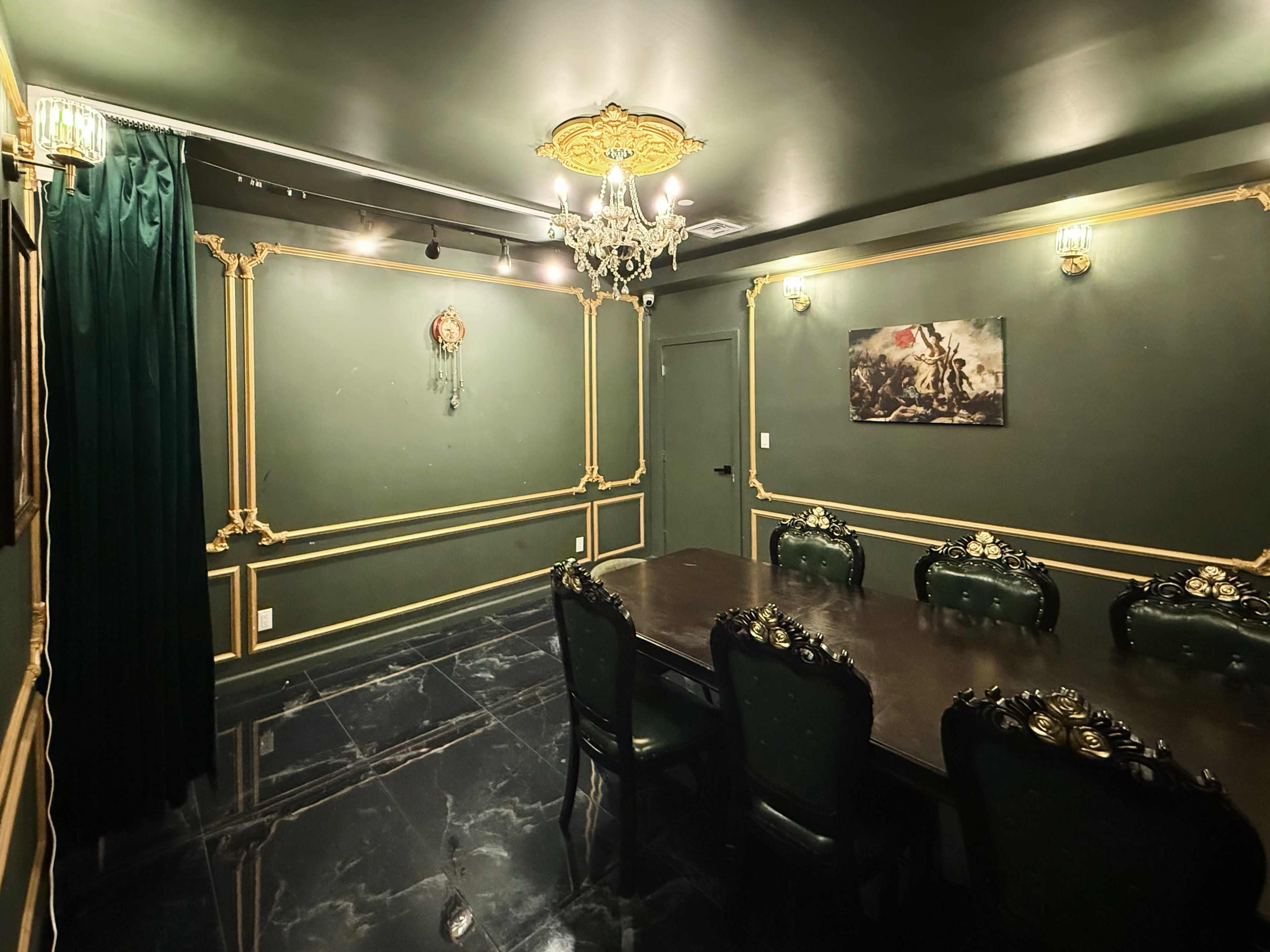The room features dark green walls with gold trim, a large dark wood table surrounded by ornate green chairs, and a chandelier hanging from the ceiling.