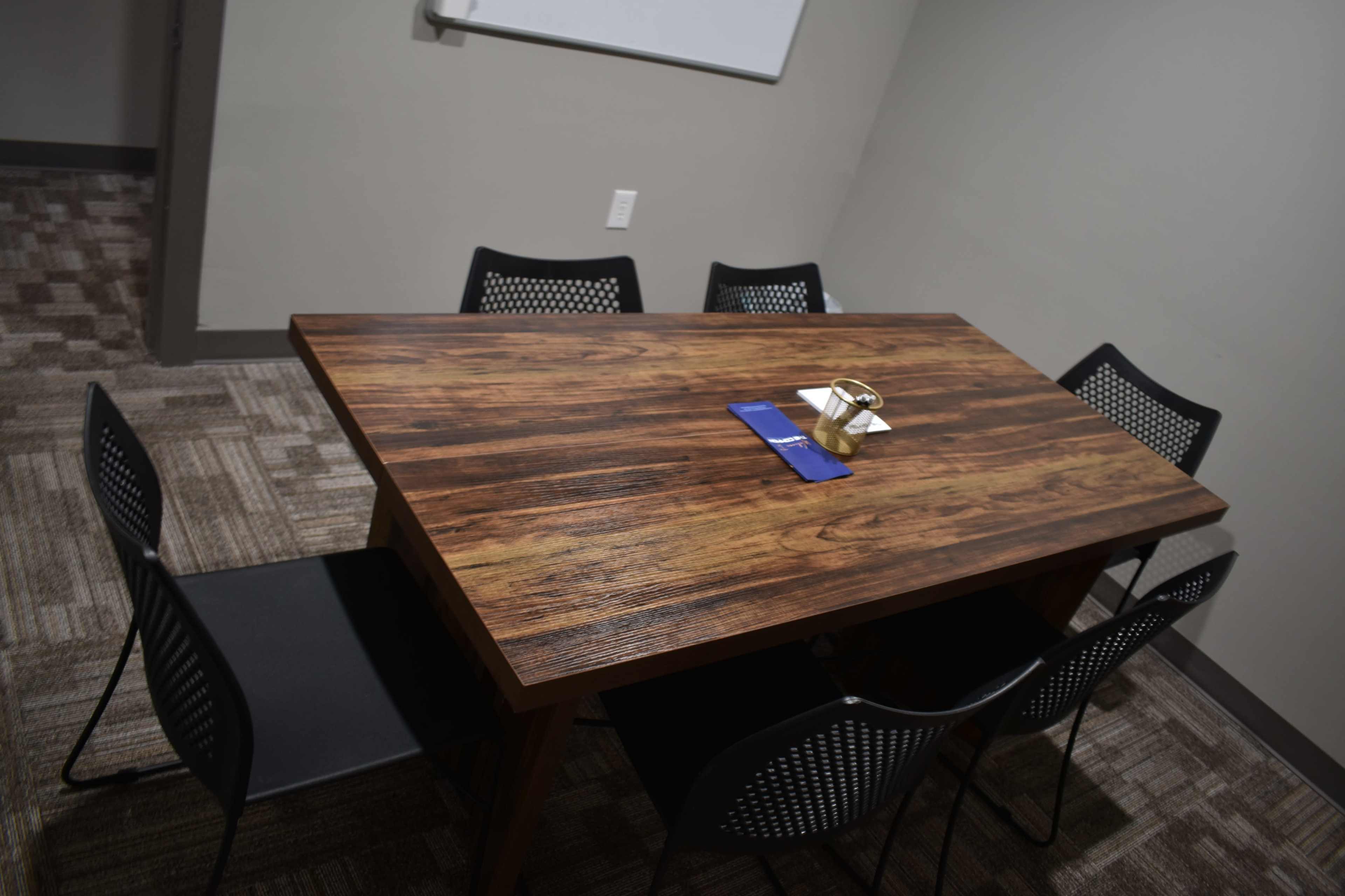 A large wooden table surrounded by six black chairs is set in a small, plain meeting room with a whiteboard in the background.