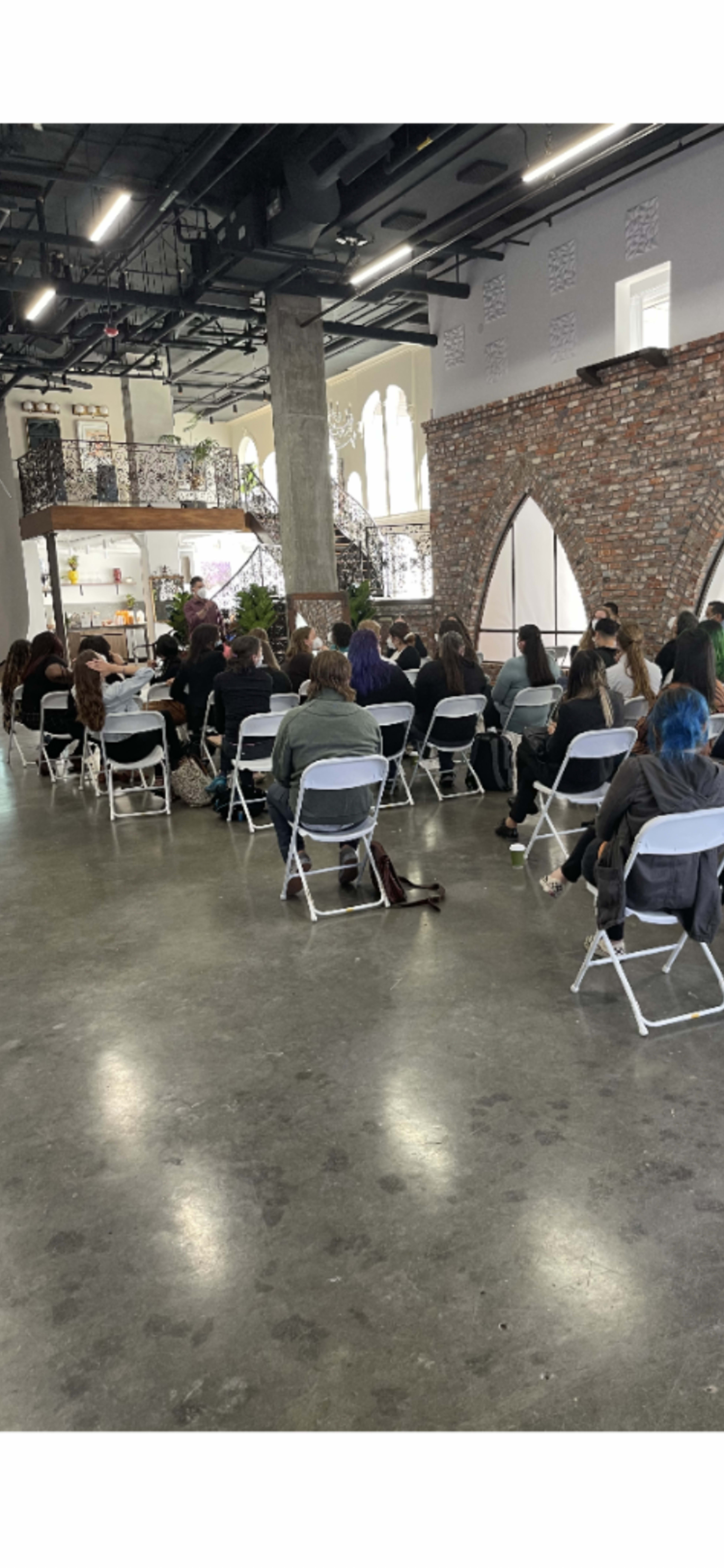 A group of people sits in rows of white chairs in a spacious interior with exposed brick walls and large windows.