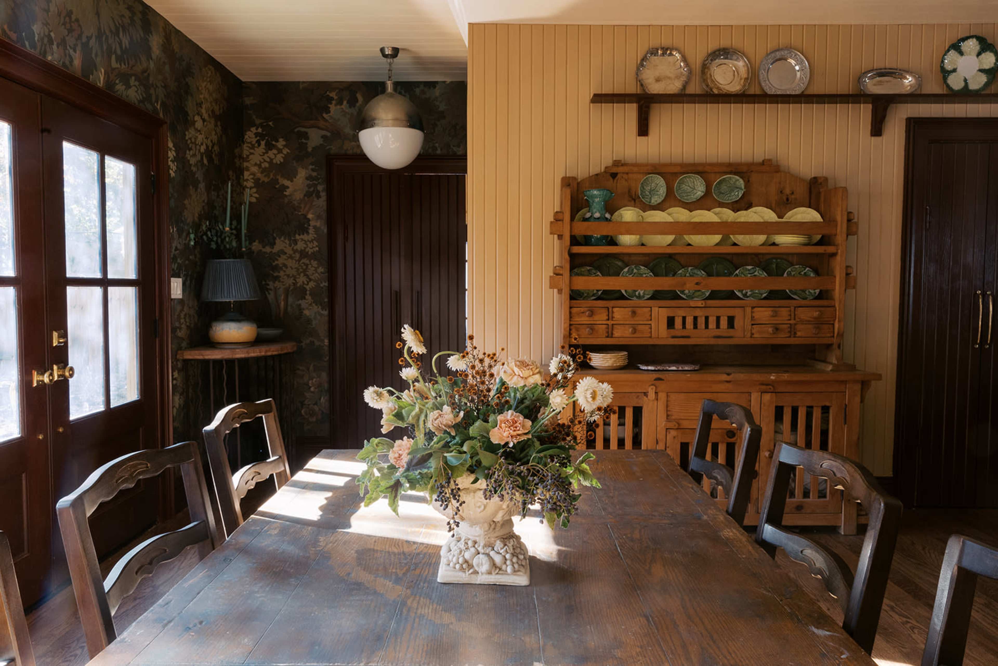A wooden dining table with a floral centerpiece is surrounded by chairs in a room featuring patterned wallpaper and a rustic wooden sideboard displaying dishware.