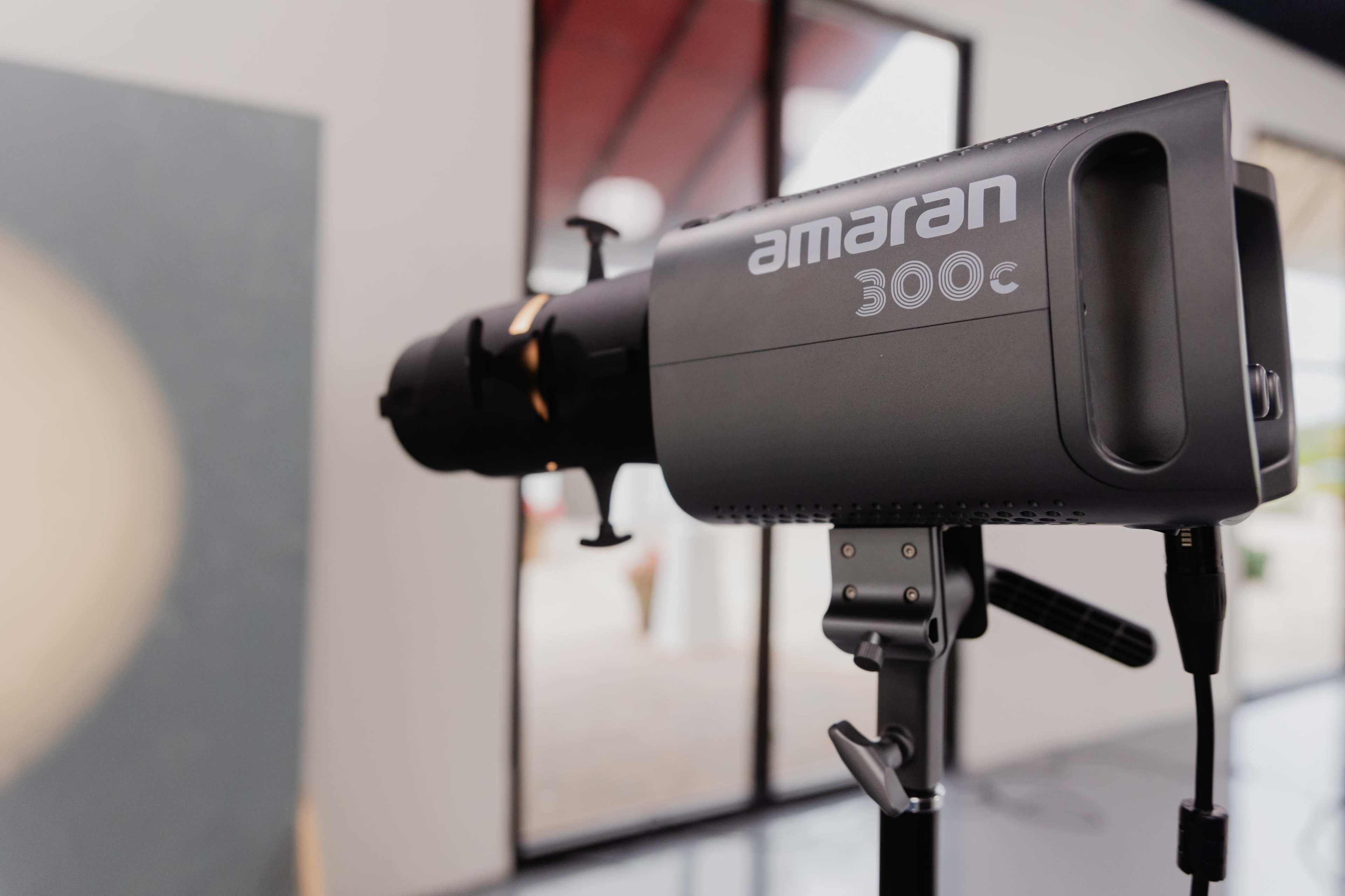 A professional lighting fixture is mounted on a stand in a studio with large windows in the background.