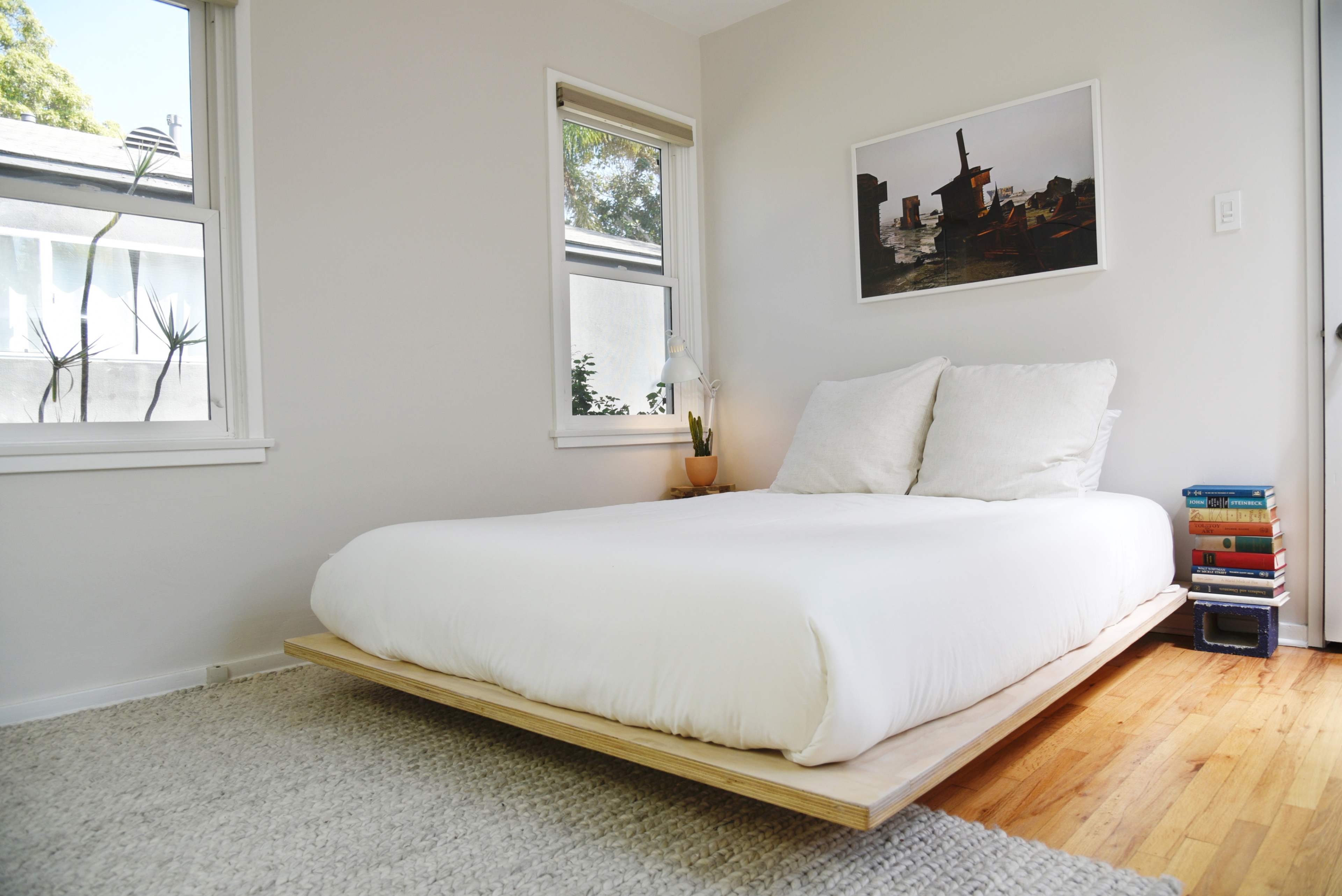 The room features a minimalist bed with a wooden frame, a small plant on a bedside table, and books stacked on the floor beside it.