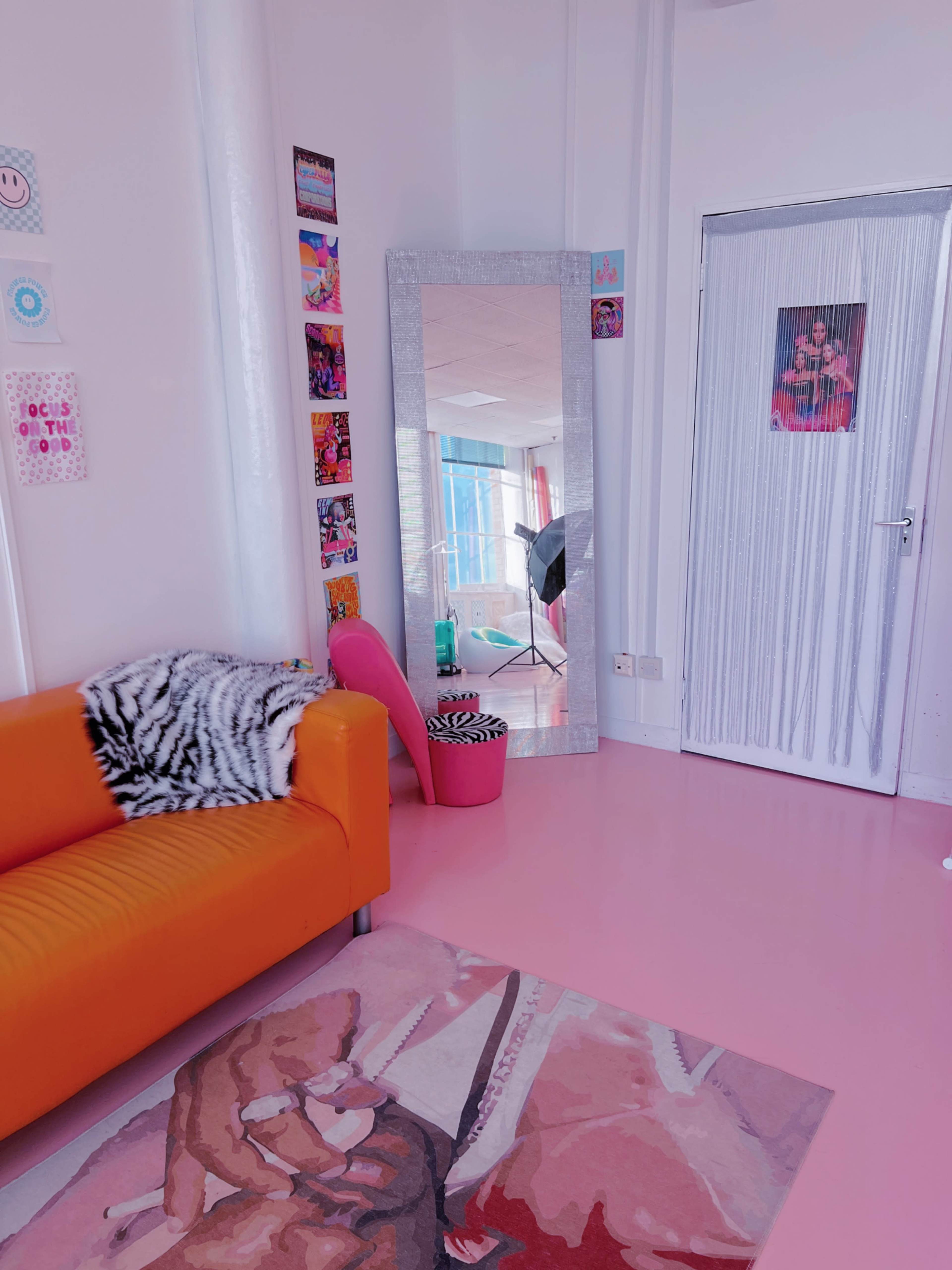 A colorful interior space featuring an orange couch, a large mirror, and a pink chair, with a light pink floor and various decorations on the walls.