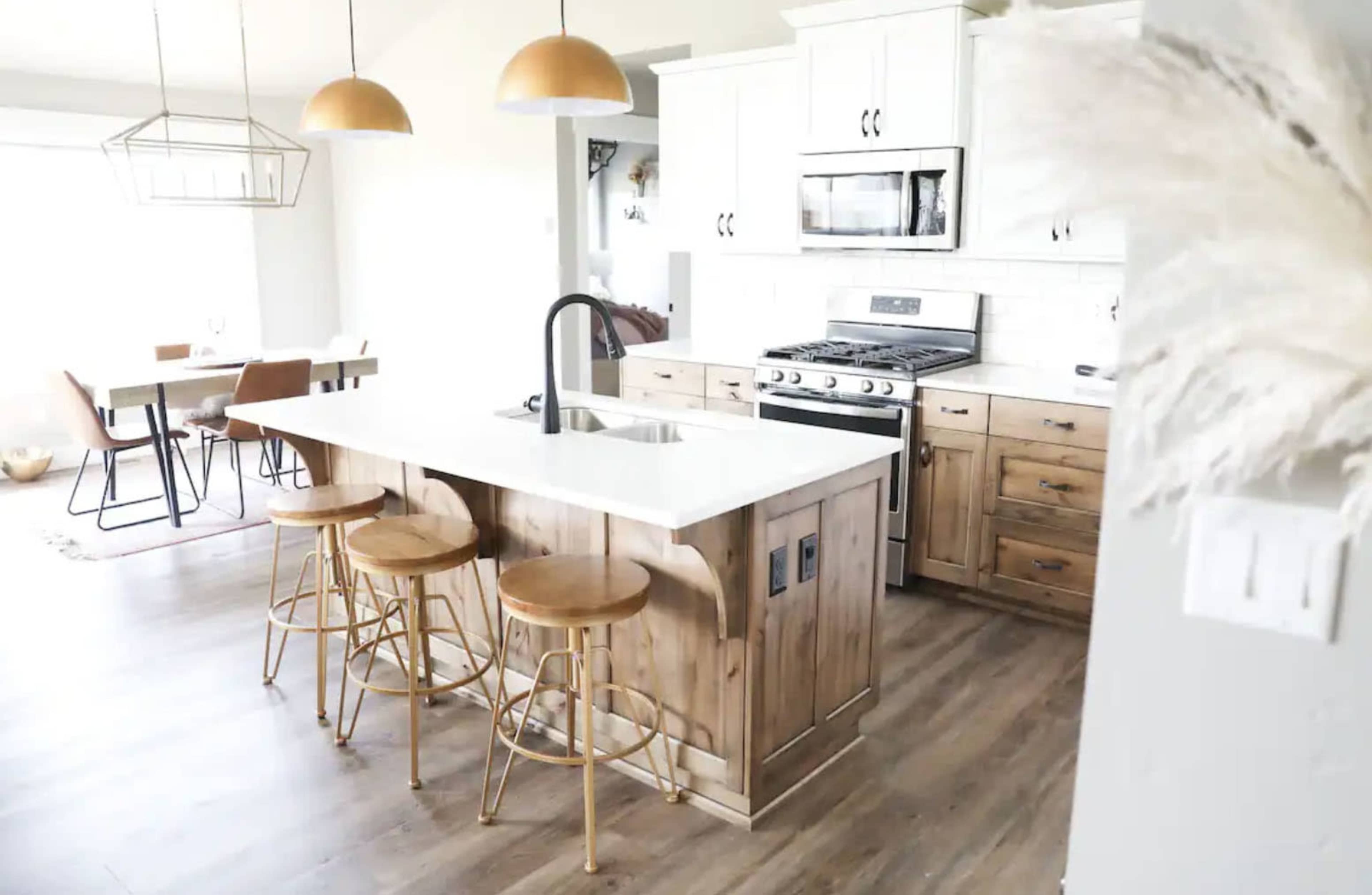 A modern kitchen features a central island with three wooden stools, equipped with a sink and surrounded by light-colored cabinets, a stainless steel stove, and large windows.