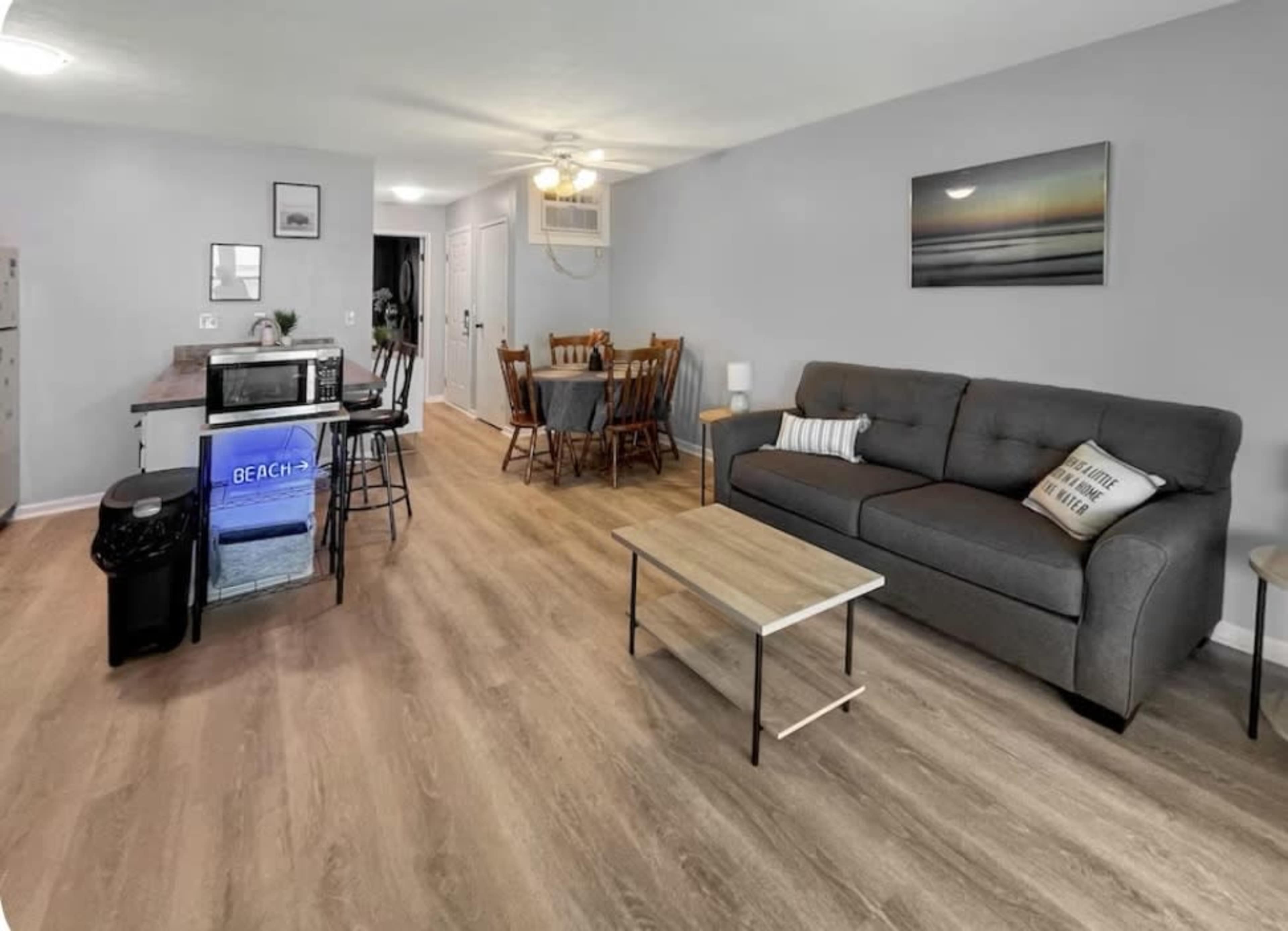 The image shows a modern living area with a gray sofa, a coffee table, a kitchenette with bar stools, and a dining table set for six.