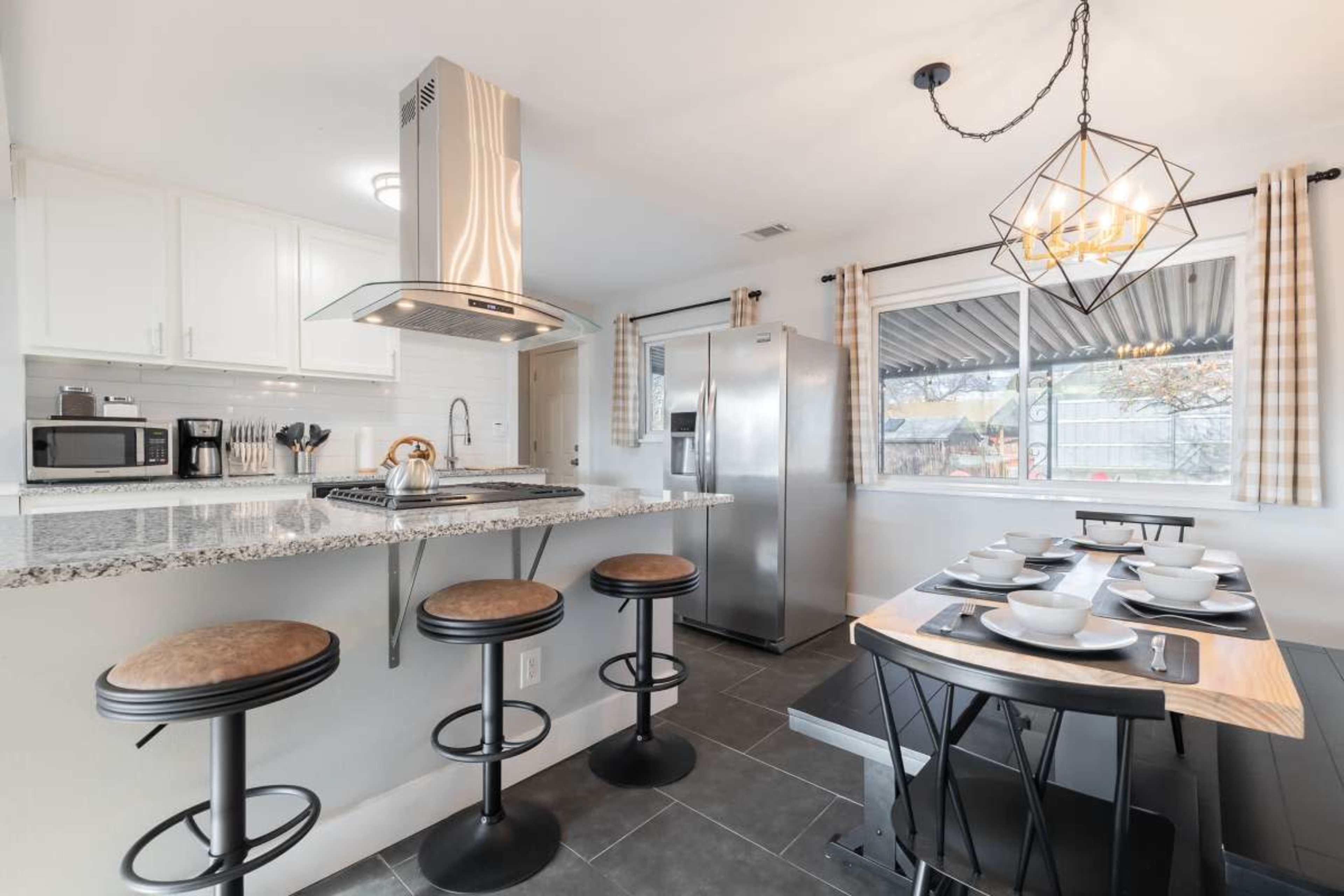 The image shows a modern kitchen with a bar counter, stools, stainless steel appliances, and a dining area featuring a table set for several people.