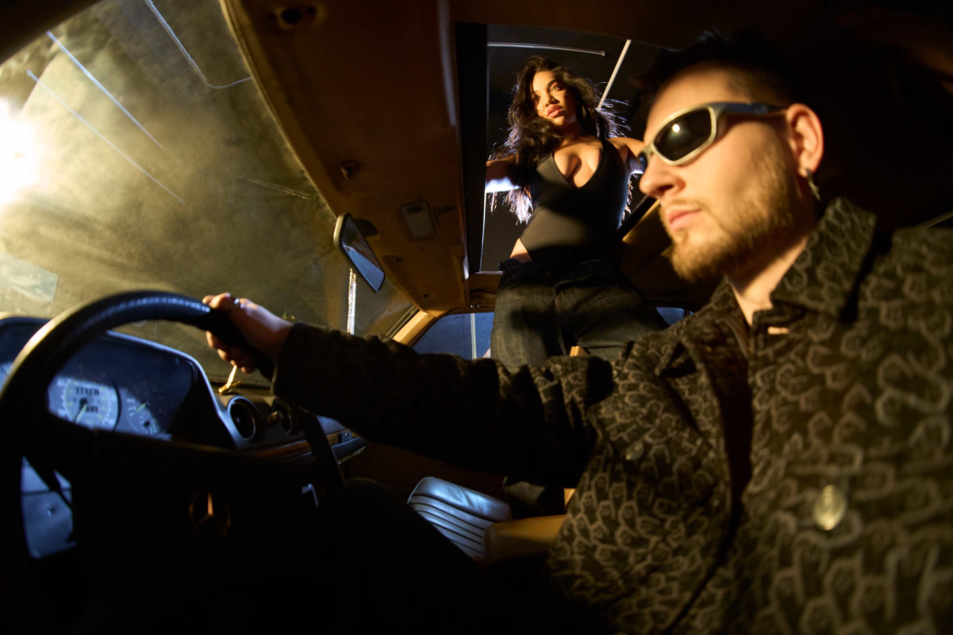 A man in sunglasses sits in the driver's seat of a car while a woman stands outside, partially visible through an open roof.