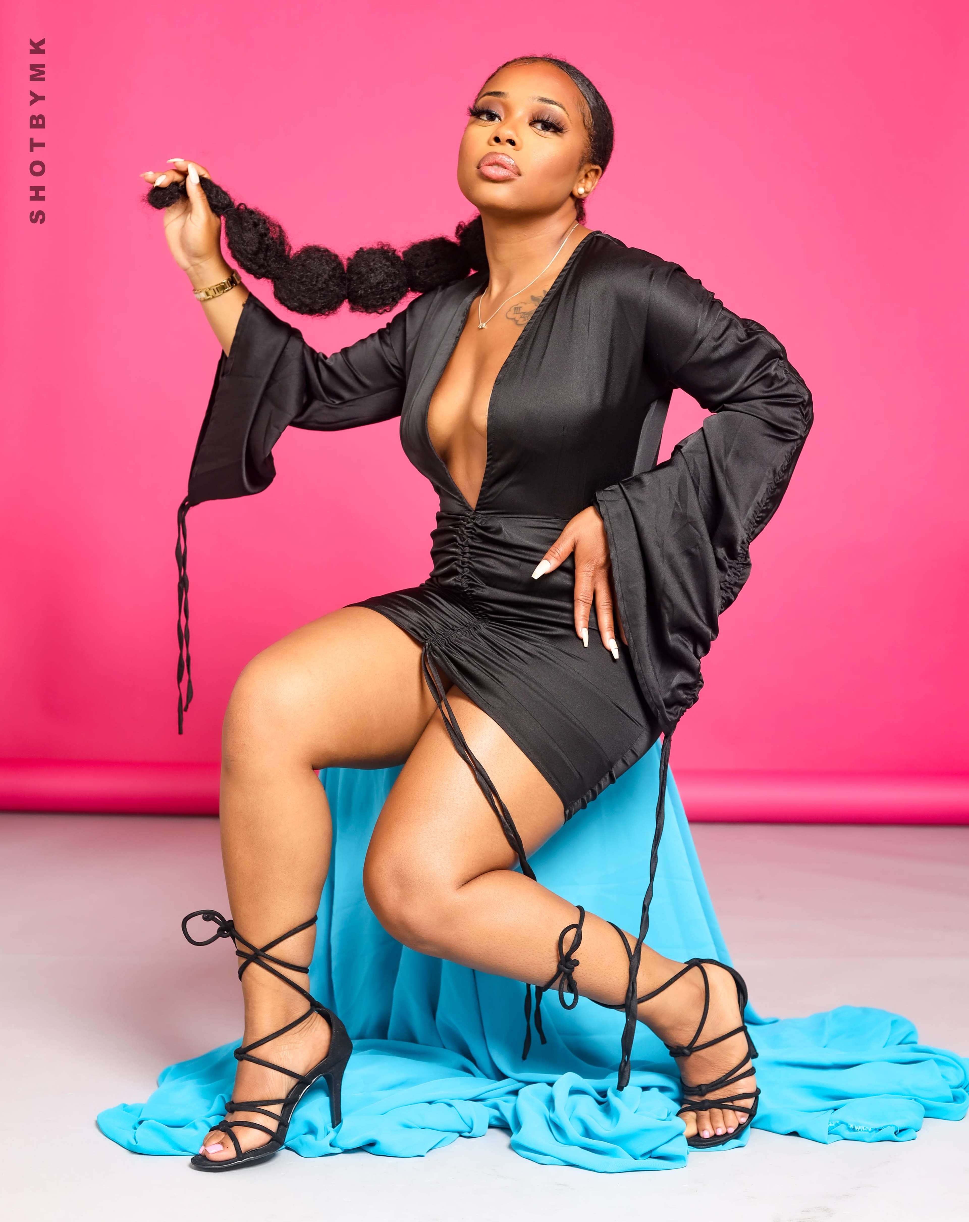 A person poses confidently in a black dress against a bright pink background, seated on a blue fabric draped on the floor.