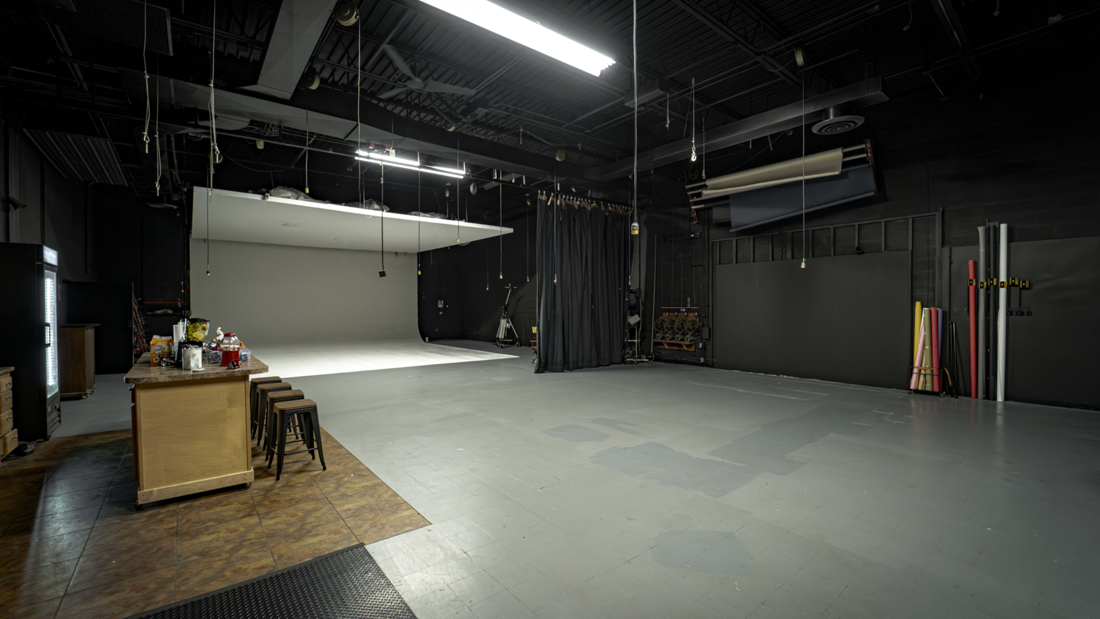 The image shows a spacious photography studio with a minimalist backdrop, equipment on the walls, and a snack preparation area on one side.