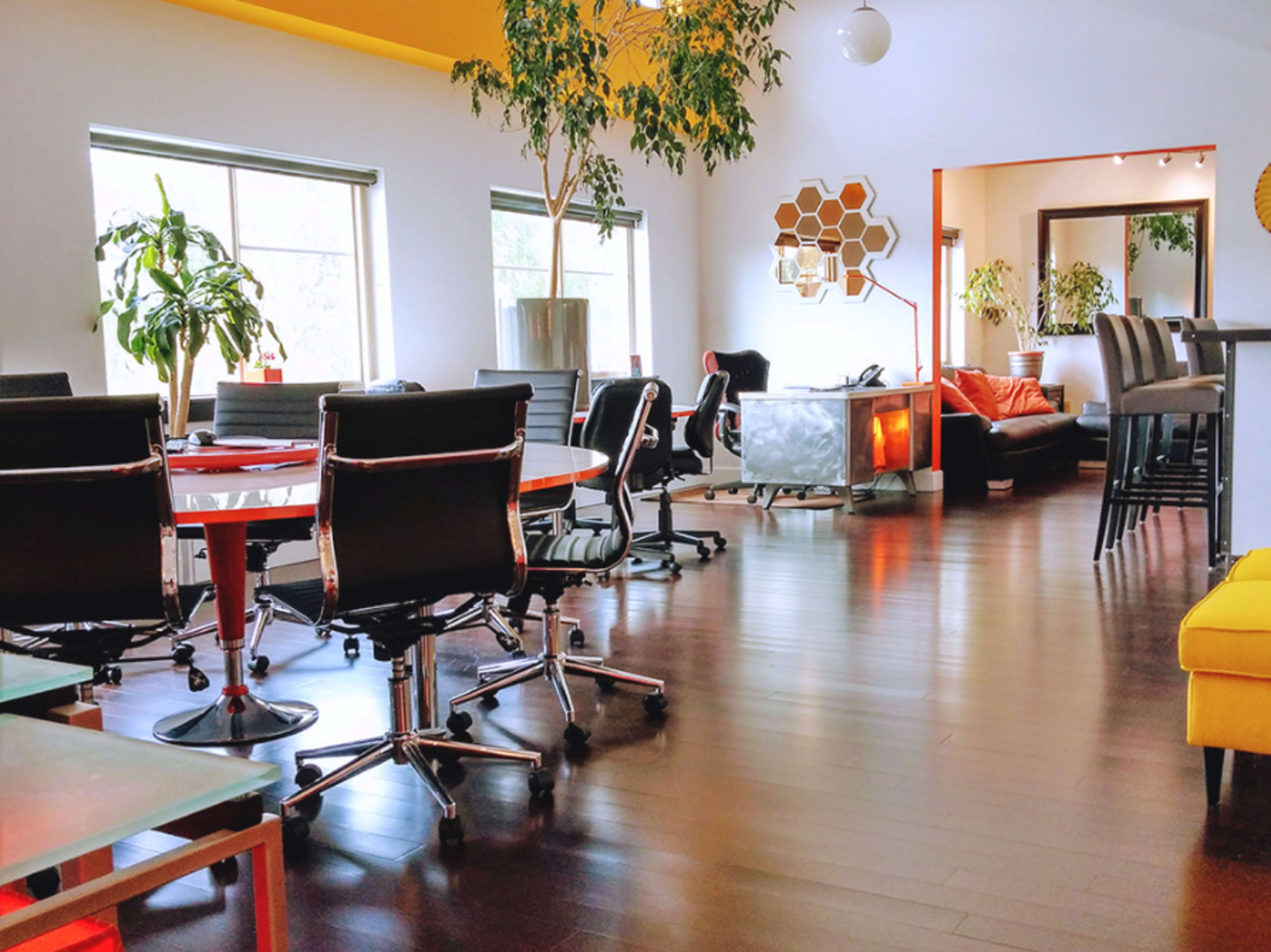 A modern office space featuring several desks and chairs, large windows, plants, and a seating area with a couch.