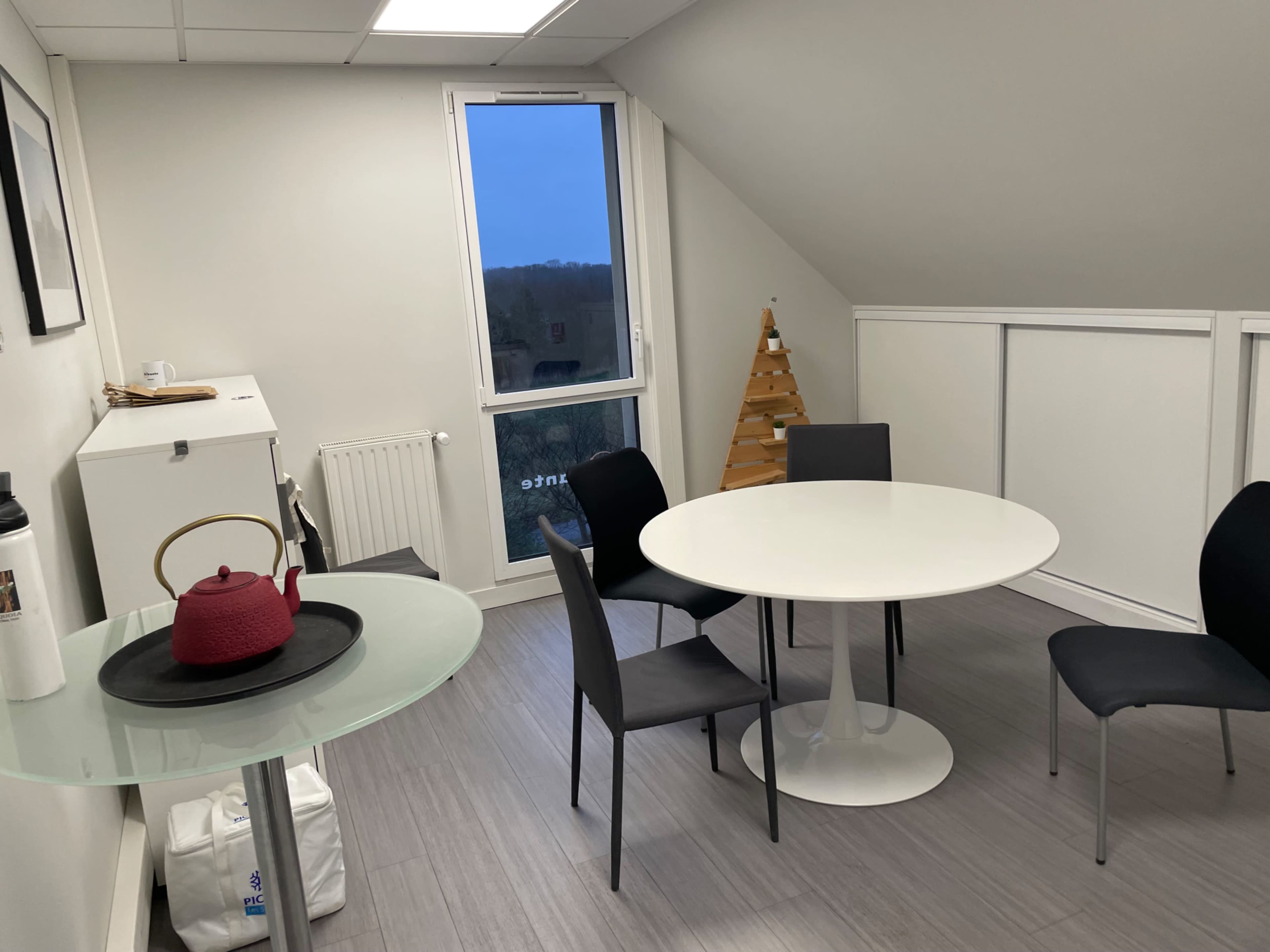 The image shows a modern, minimalist meeting room with two tables, several chairs, and a small decorative tree in the corner.