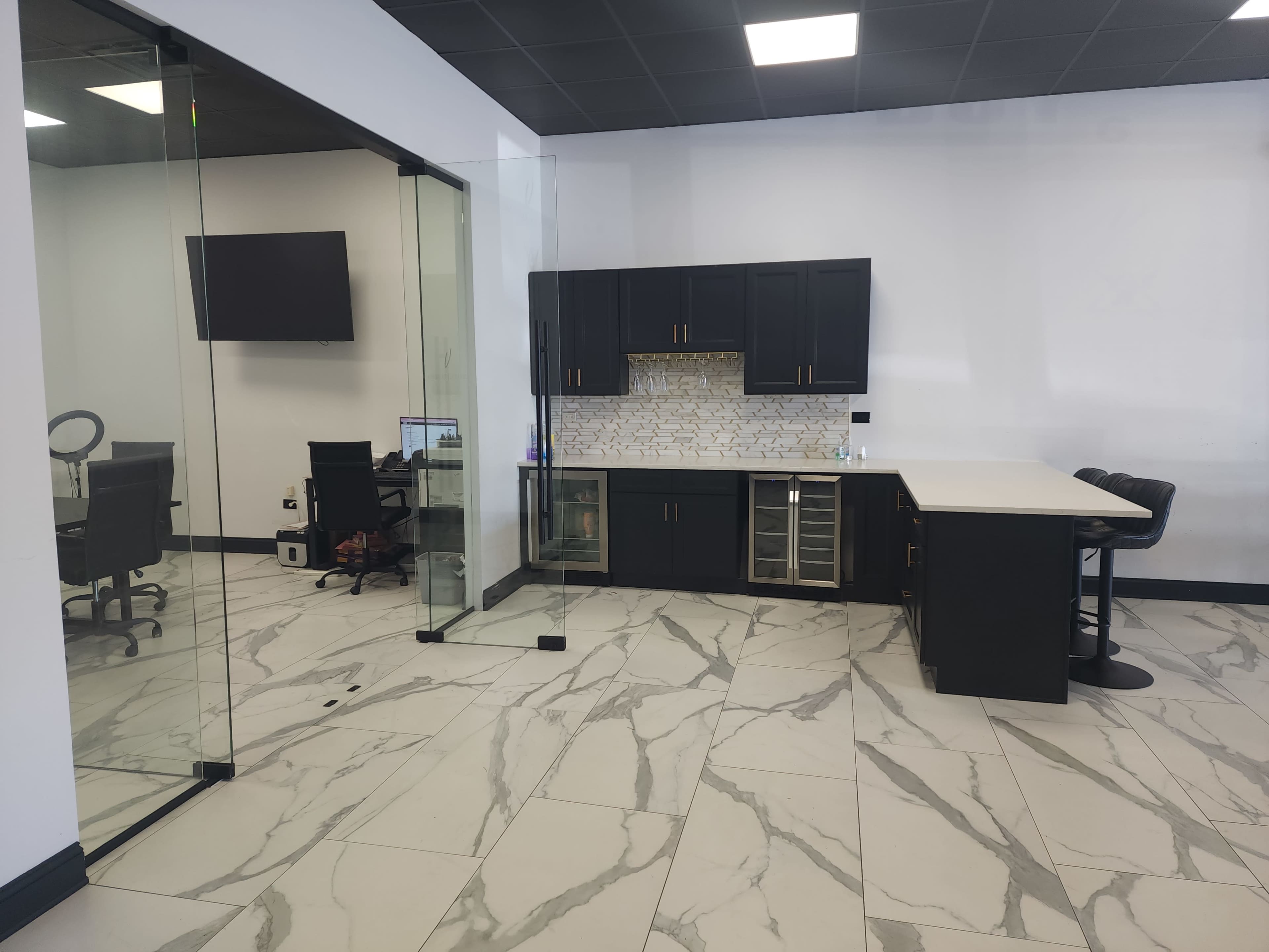 The image shows a modern office space featuring a glass partition, a kitchenette with black cabinets, and a seating area with bar stools.