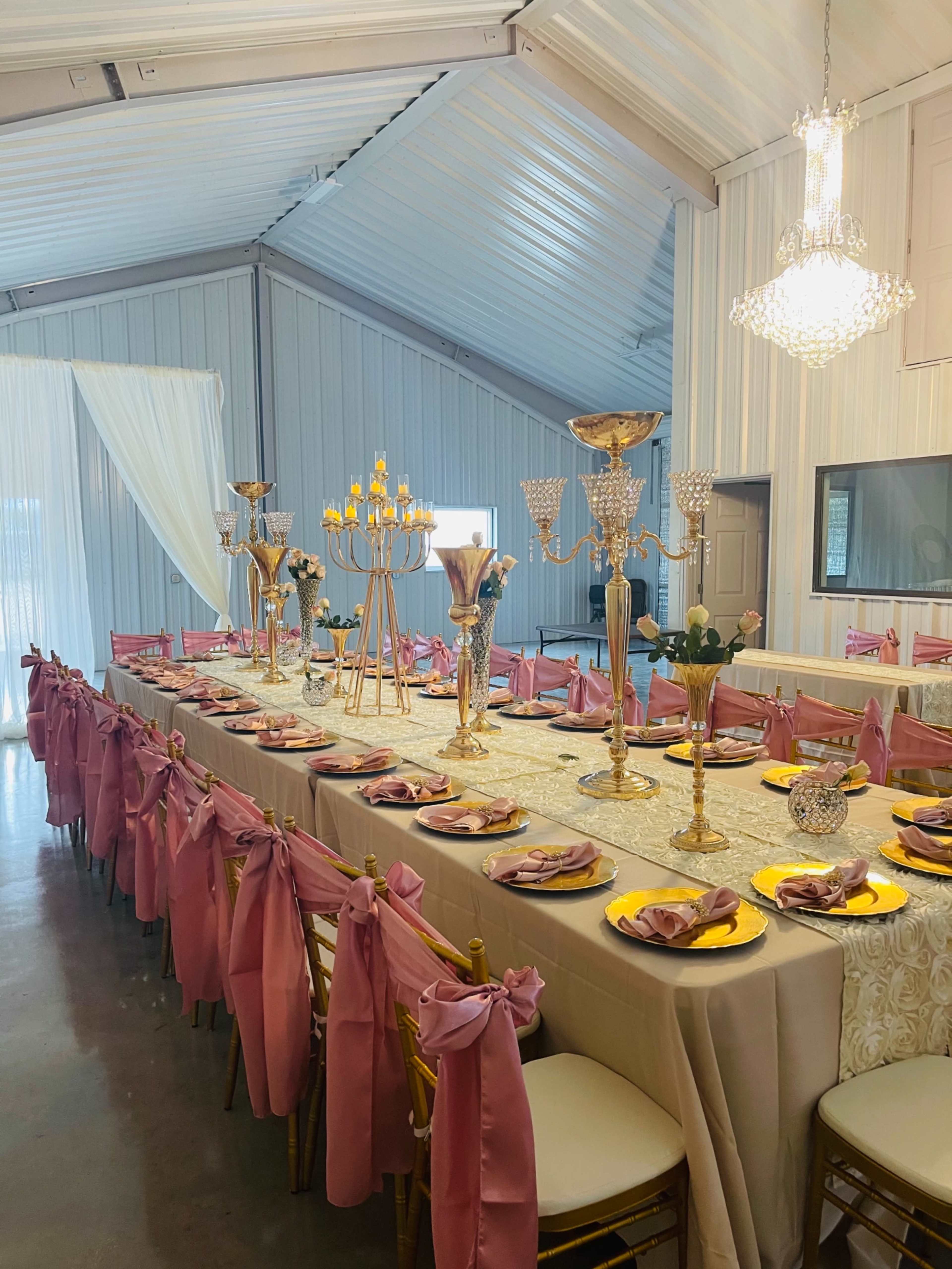 A long banquet table is elegantly set with pink napkins, golden utensils, and decorative centerpieces under a chandelier in a light-colored venue.