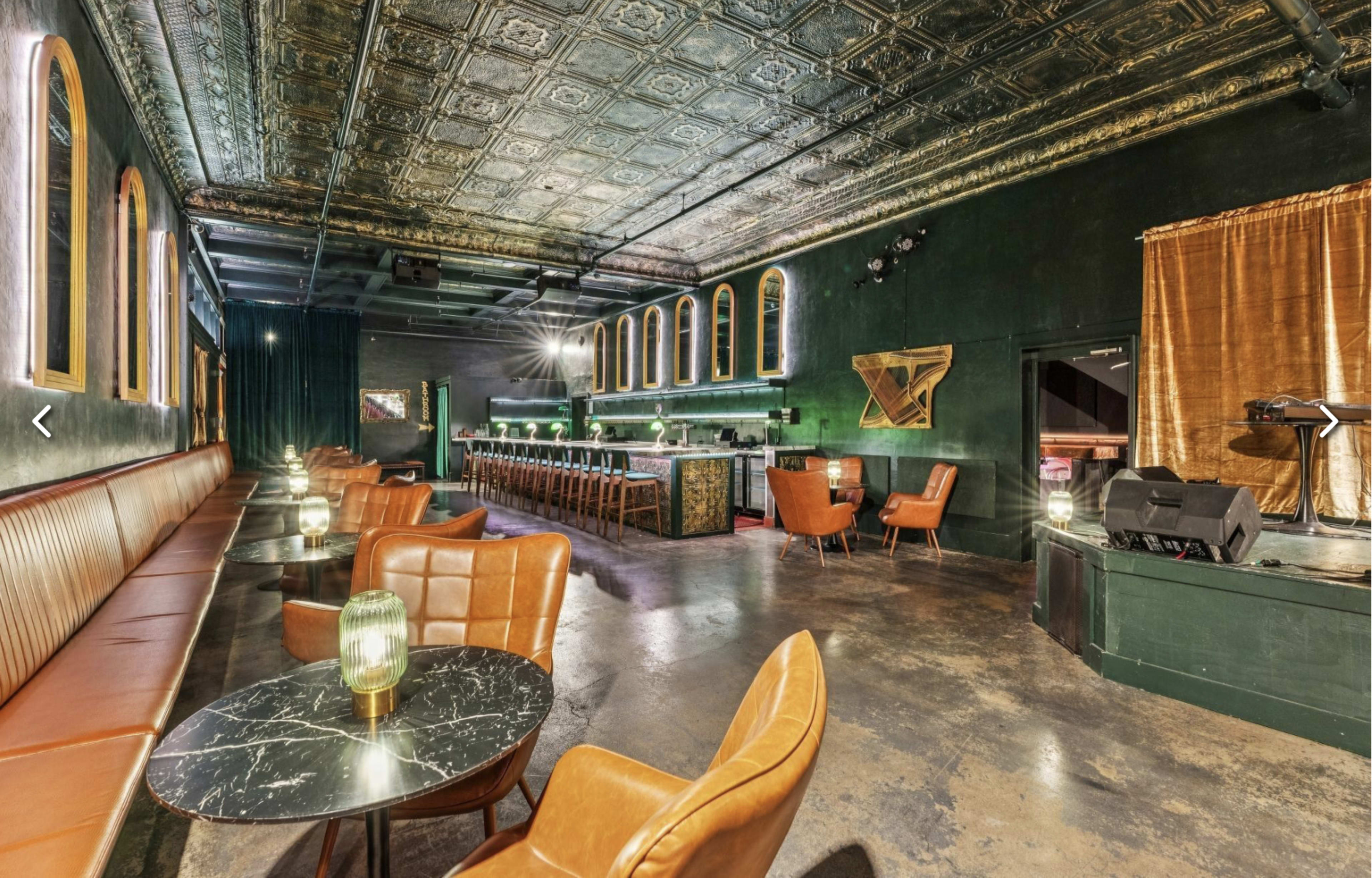 The image shows a dimly lit bar with dark green walls, vintage-style seating, and a long bar counter illuminated by stylish lighting fixtures.