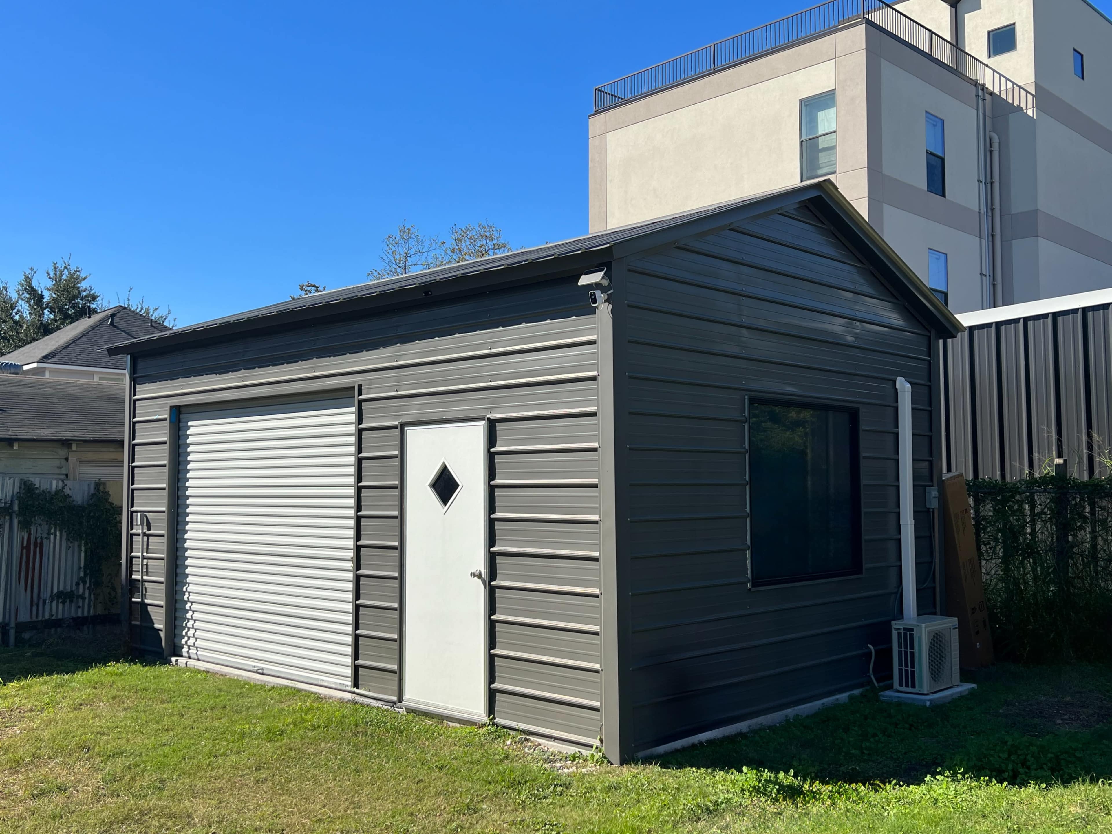 A metal storage shed with a roll-up door and a side entrance is situated in a grassy area next to a tall building.