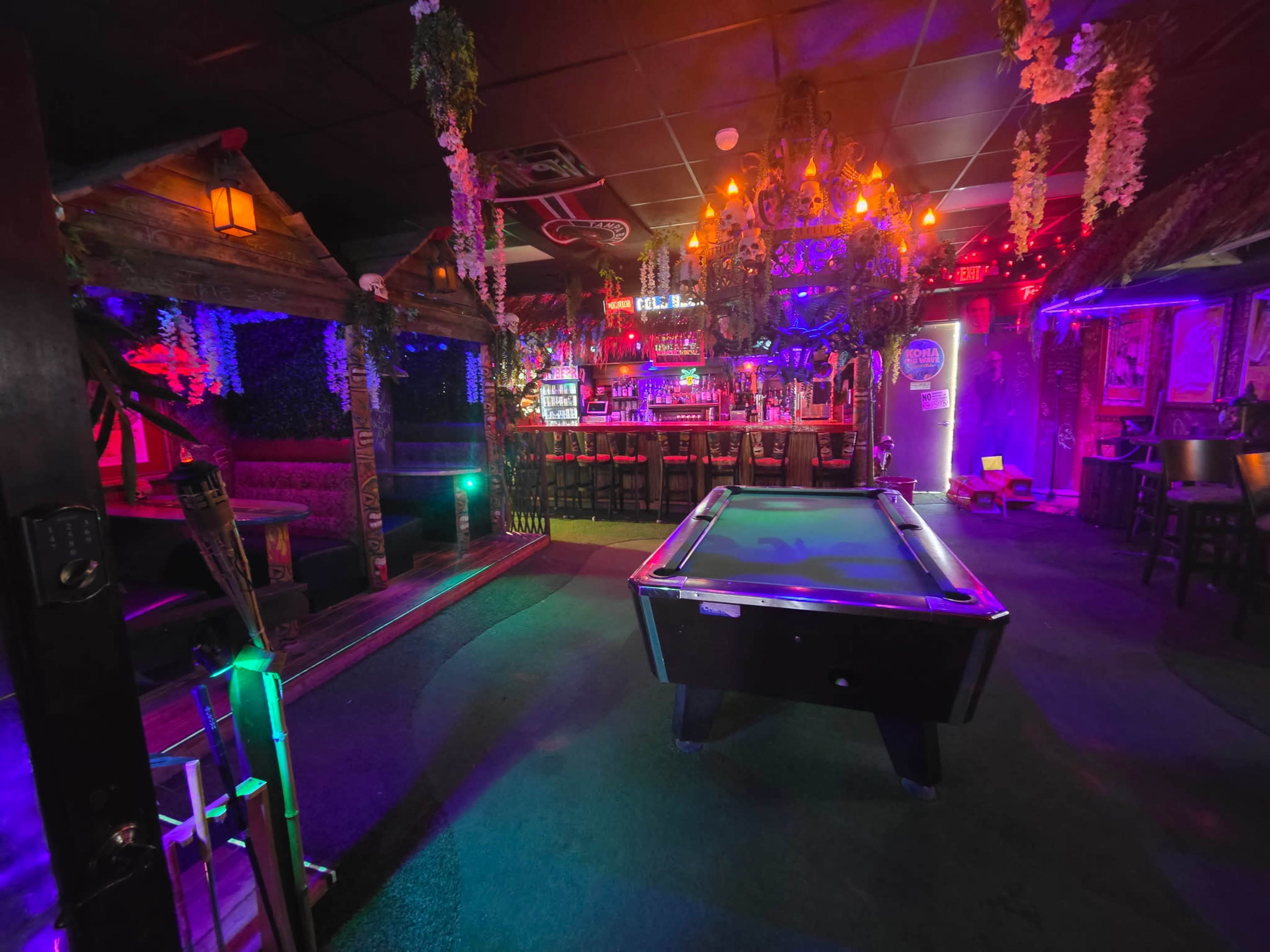 The image shows a vibrant lounge area with a pool table at the center, surrounded by cozy seating and colorful lighting.