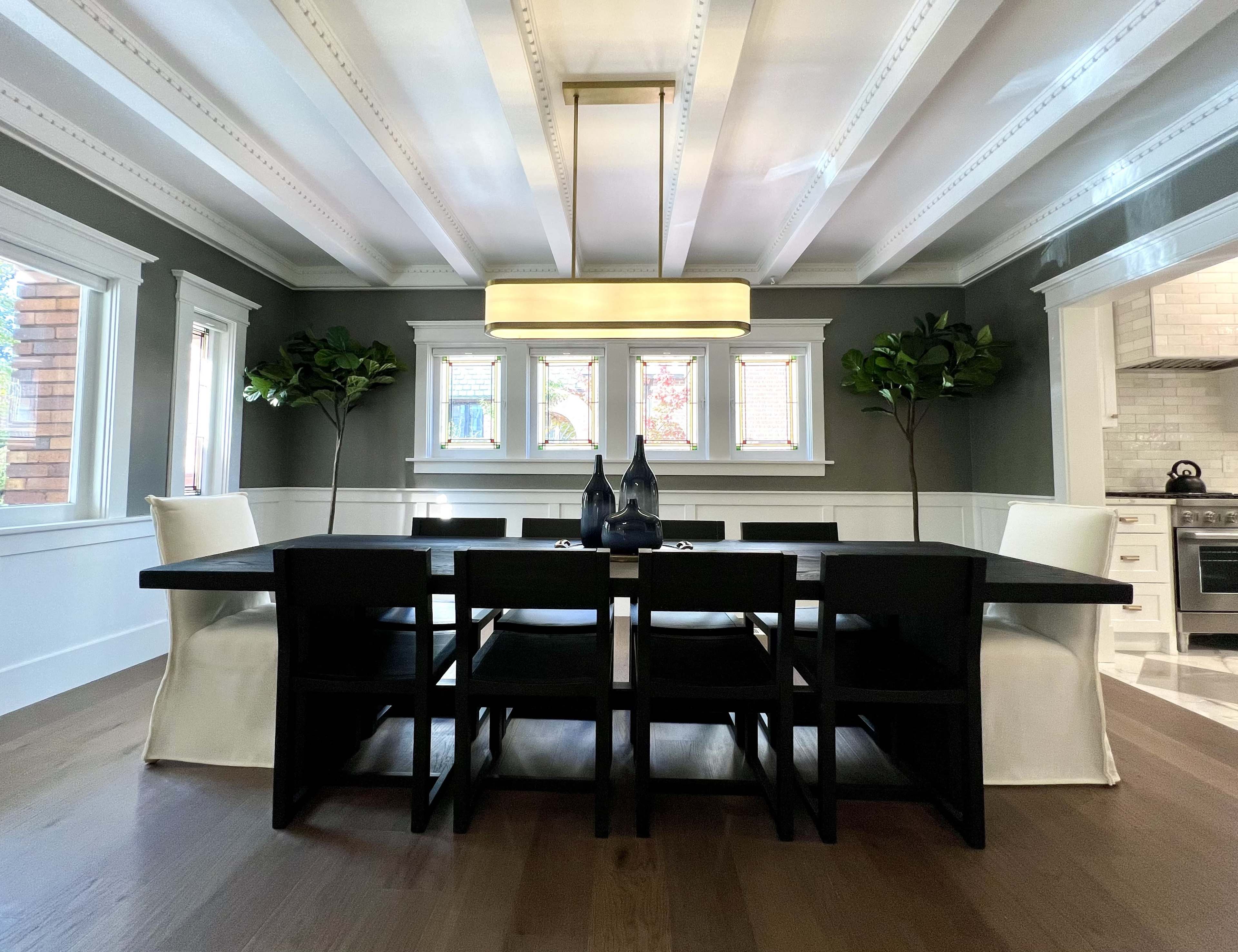 A modern dining room features a black table and chairs with white upholstered accents, illuminated by a large ceiling light and flanked by potted plants near bright windows.