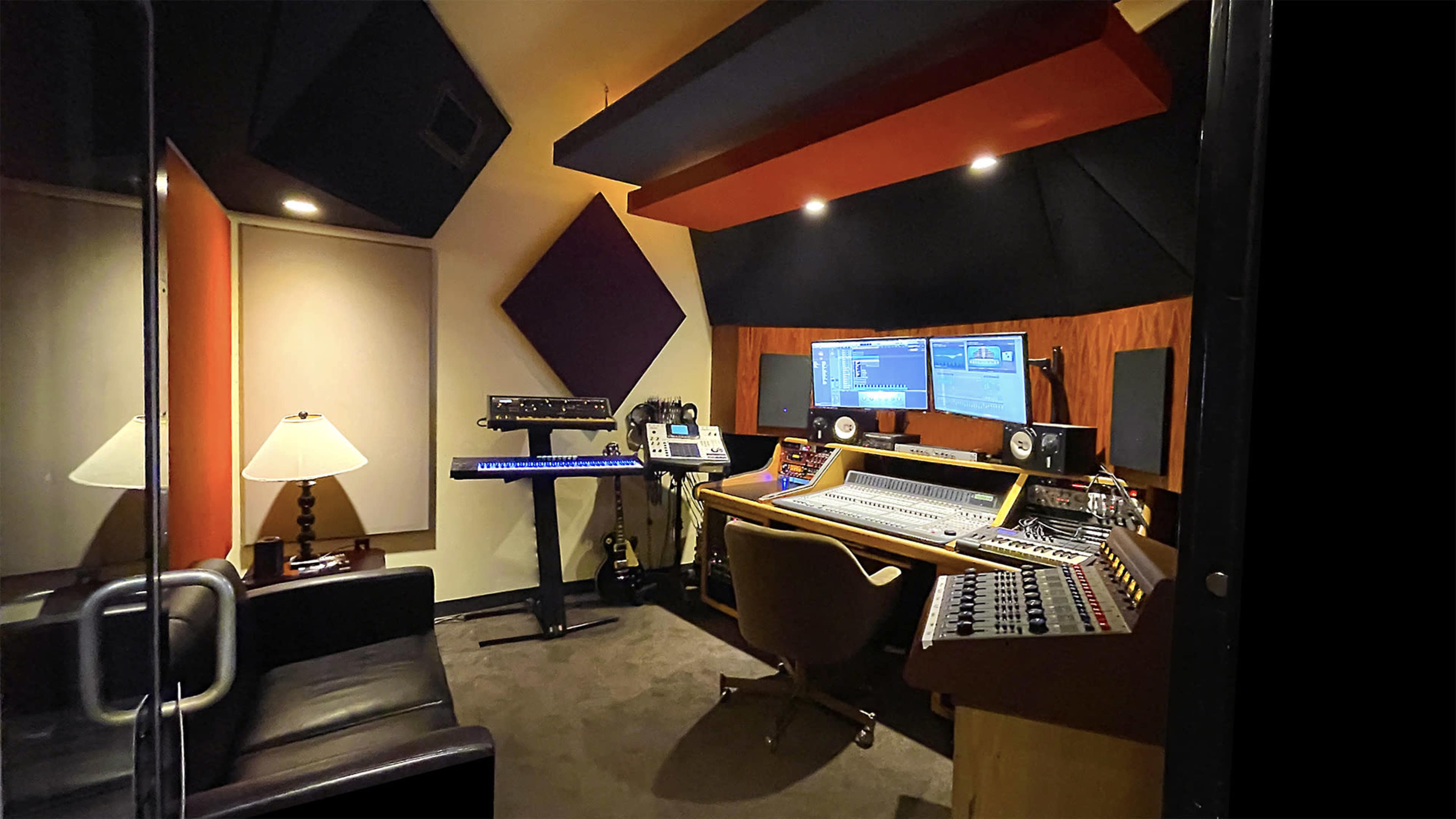 The image shows a music studio with two monitors, a mixing console, and various audio equipment, including a keyboard and a lamp.