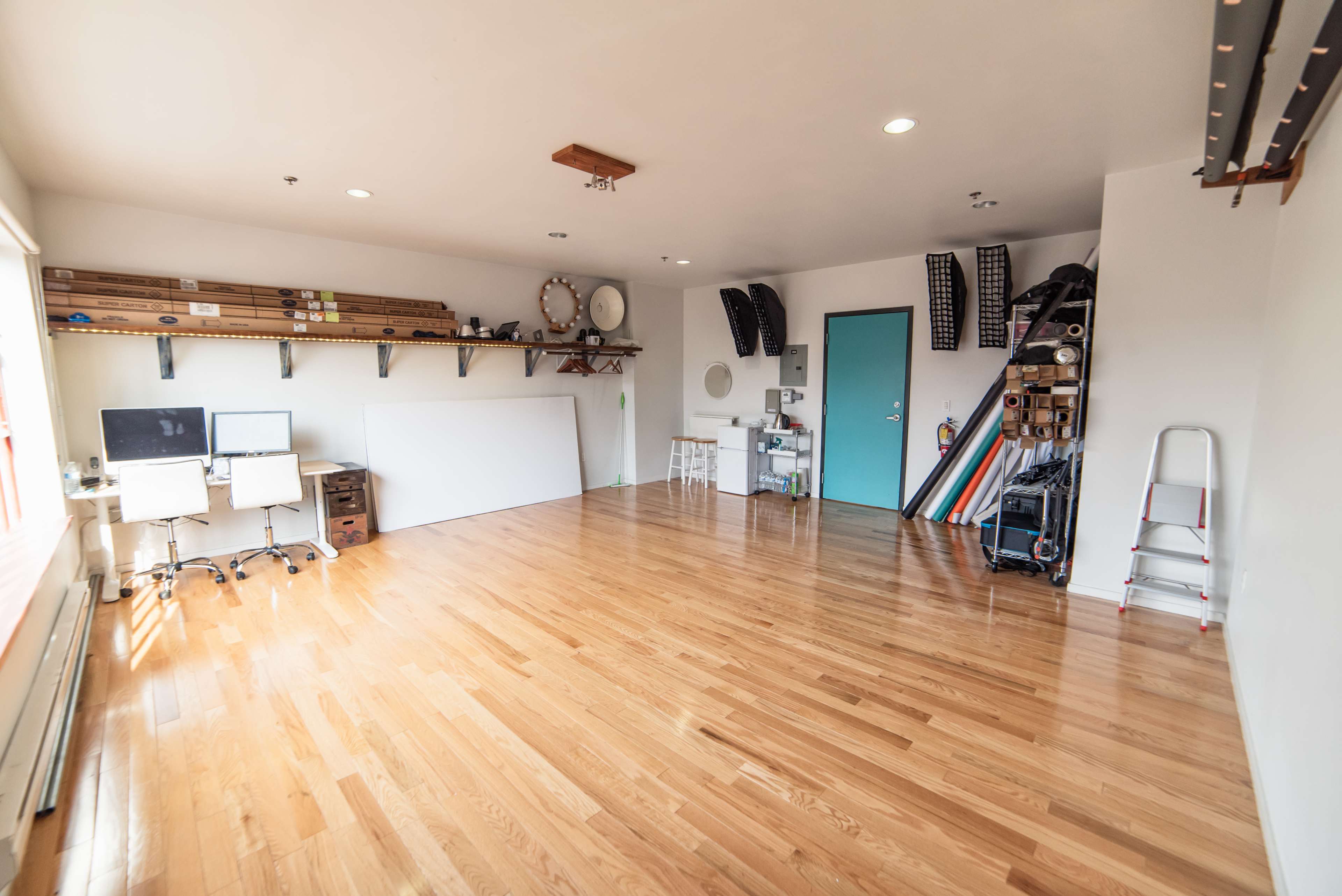 A spacious, empty room with wooden floors, featuring two workstations with computers, shelving along the walls, and various equipment stored in the corner.