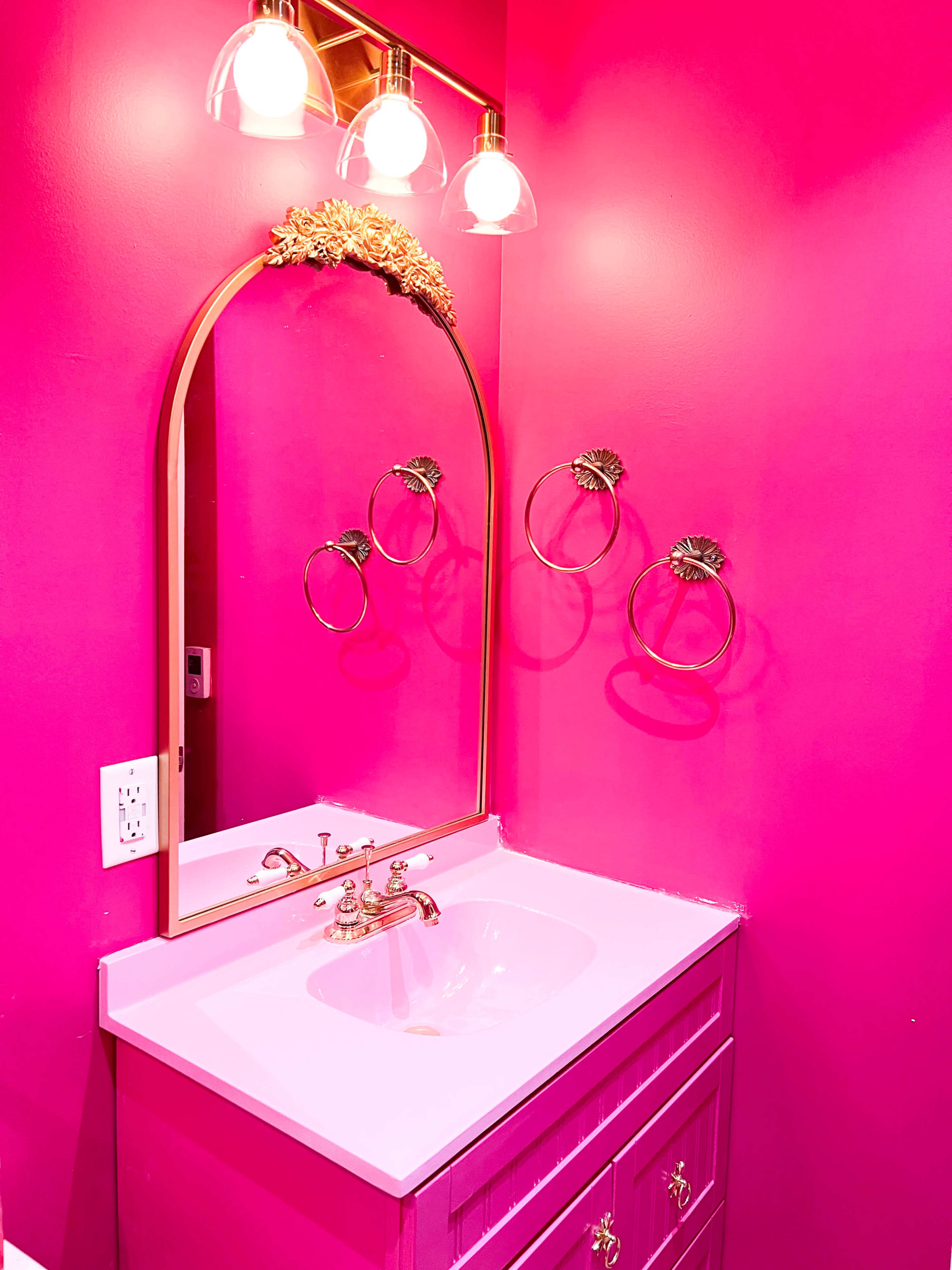 A bright pink bathroom featuring a sink with a mirror above it and three decorative towel rings on the wall.