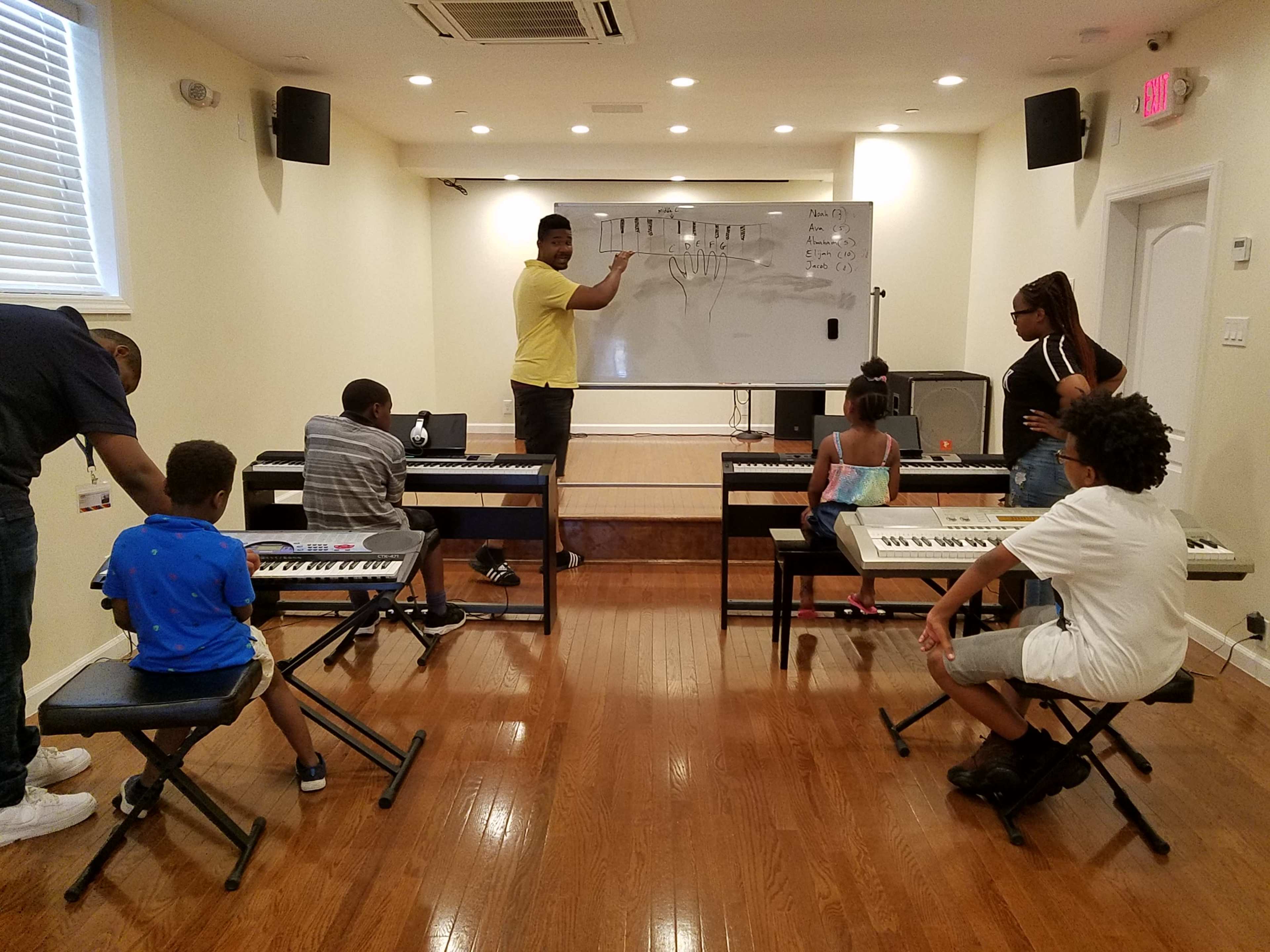 A music instructor stands in front of a whiteboard while several students sit at keyboards in a classroom setting.