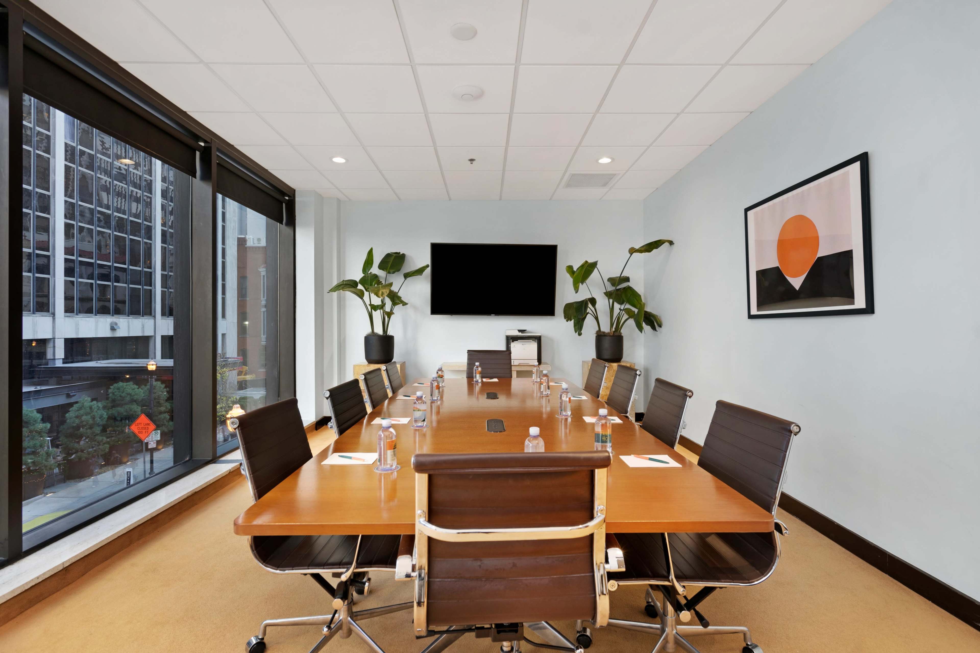 A modern conference room with a long wooden table, black leather chairs, a wall-mounted TV, and large windows overlooking a city street.