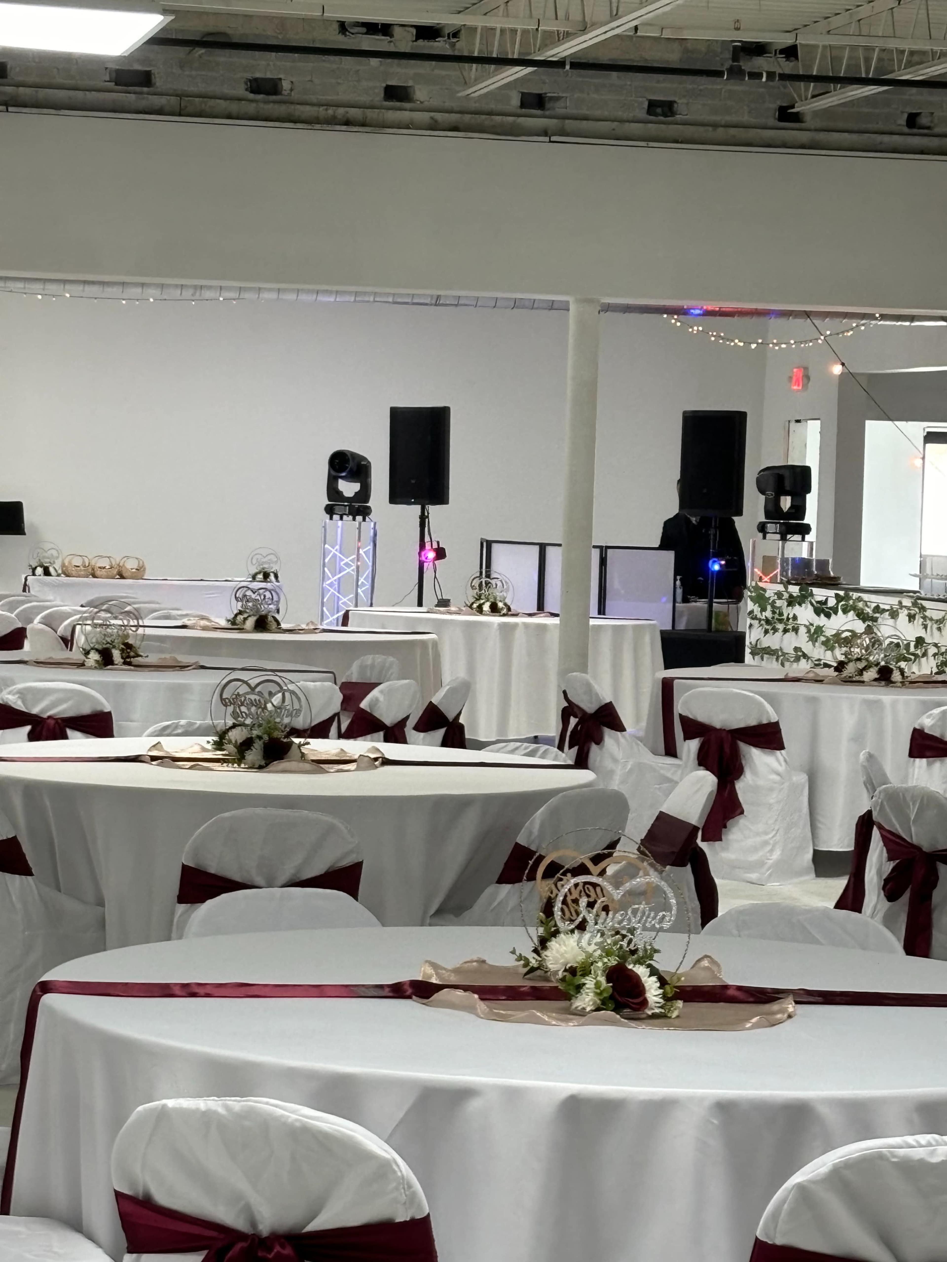 A banquet hall set up for an event, featuring round tables with white tablecloths and burgundy sashes, along with decorative centerpieces and audio equipment in the background.